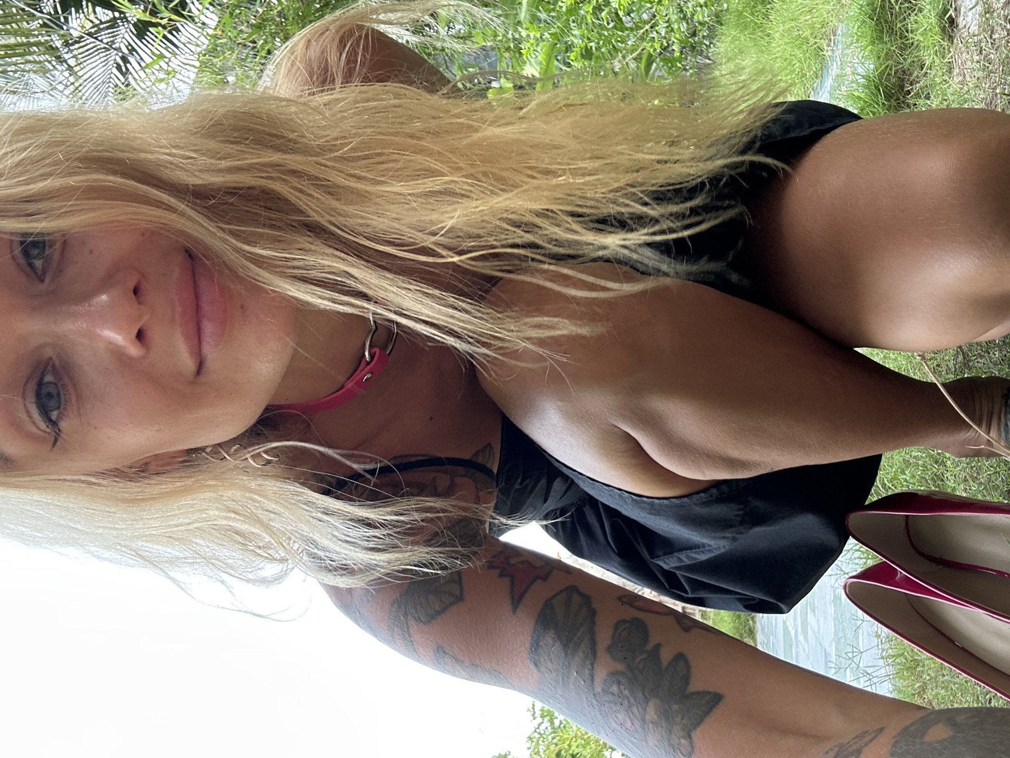 A blonde woman, positioned low to the ground with her knees bent, is the central subject. She wears a black dress and a pink choker necklace. Her left arm, covered in tattoos, is visible, as is her right thigh. In the foreground, a pair of shiny pink high-heeled shoes rests on the grass. The background consists of lush green foliage and a body of water, likely a river or canal, suggesting a natural or semi-rural environment. The scene is set outdoors, with a bright sky overhead. The woman is looking towards the camera with a subtle smile. This image could have been taken in the vicinity of Mũi Né, Vietnam, known for its natural beauty and diverse landscapes.
