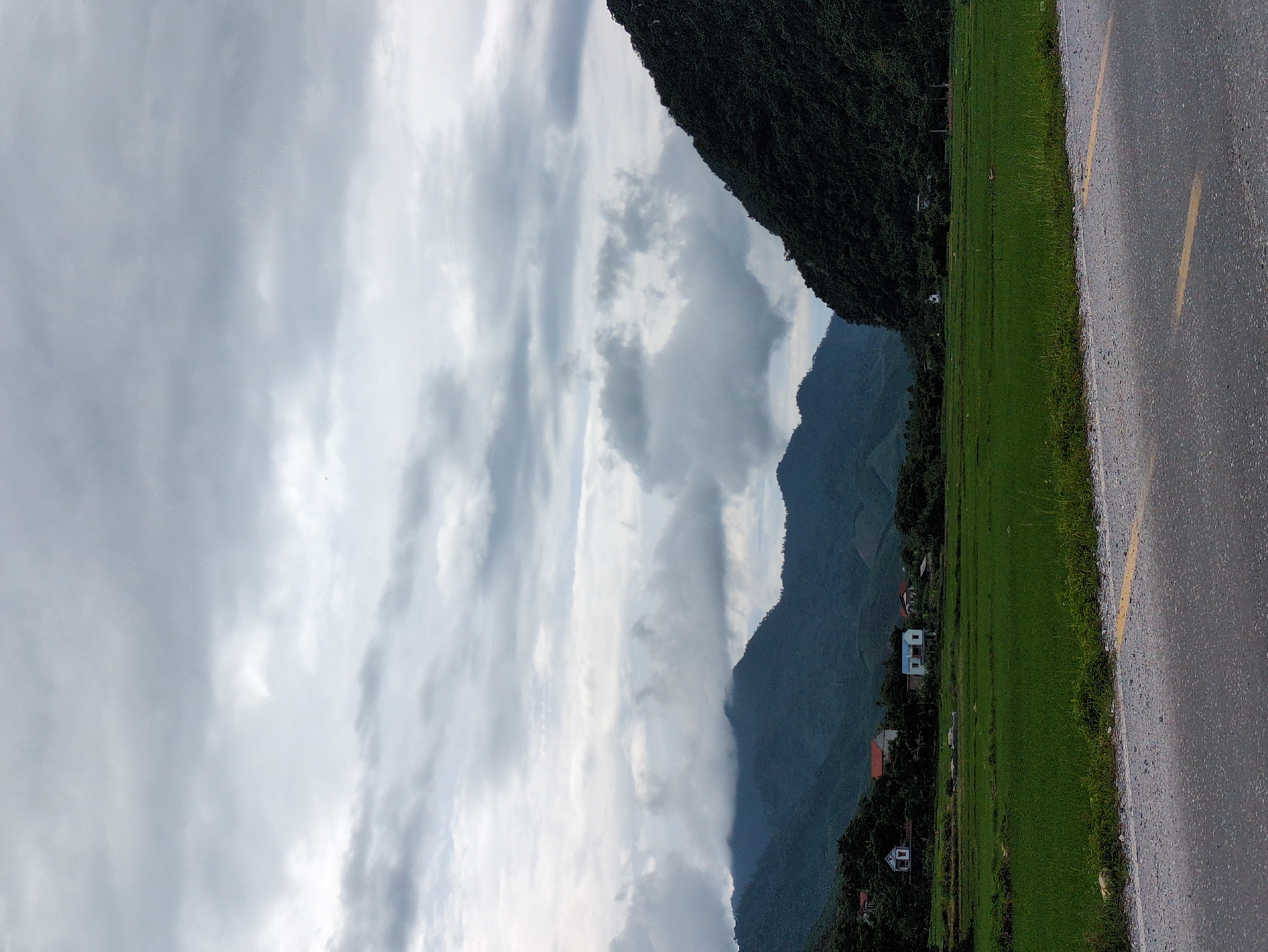 The image depicts a rural landscape under a cloudy sky. In the foreground, a gray asphalt road with a yellow line runs diagonally across the bottom of the frame. To the left of the road is a strip of green grass and some sparse vegetation. Beyond this, a vibrant green field, likely a rice paddy, stretches towards a line of trees and houses. Several houses with red and white roofs are visible nestled amongst the trees at the base of rolling, tree-covered mountains. The mountains are dark green and shrouded in mist and clouds, giving the scene a somewhat moody atmosphere. The sky is overcast with shades of gray and white clouds. The image appears to have been taken in a rural area, possibly in Tân Thanh Commune, Vietnam, given the characteristic terraced fields and mountainous terrain. There are no people or animals visible in the scene.