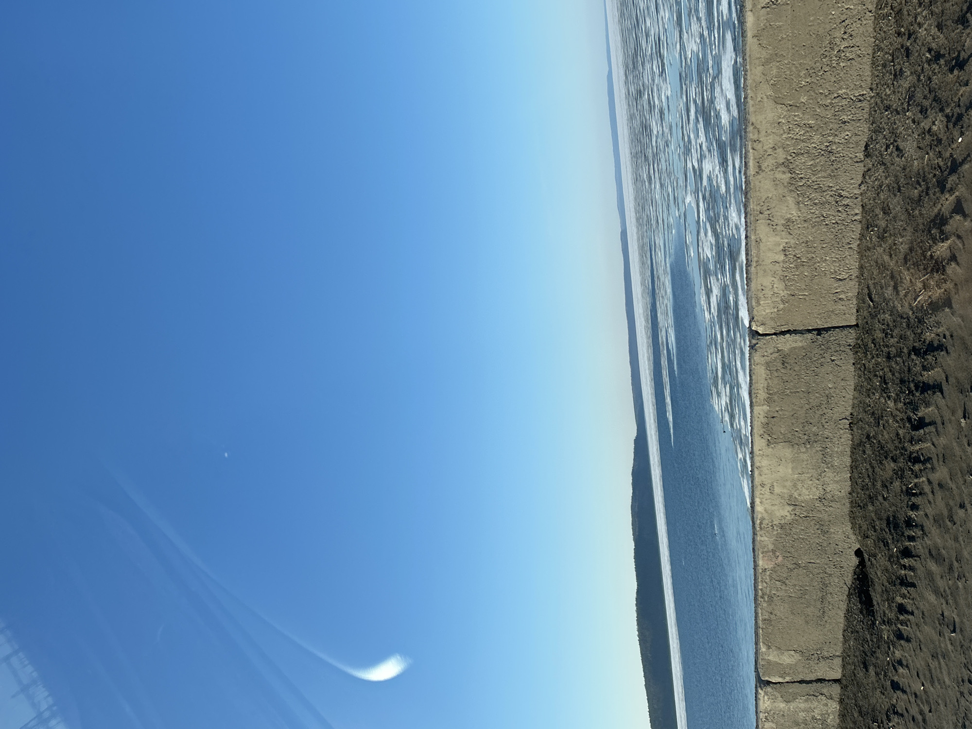 The image shows a view of the Kama River in Chaikovsky, Russia. The river is partially frozen, with ice floes visible on the water's surface. On the right side of the image, there is a concrete embankment and sandy shore. The sky is clear and blue. There are no people visible in the image.