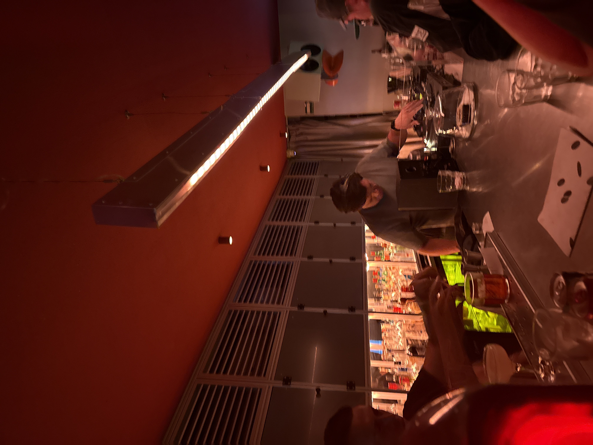 The image depicts the interior of a bar in Mexico City, Mexico. Several individuals are positioned around a bar counter, which has drinks, glasses, and a notepad with what appears to be coins. A bartender is interacting with the patrons. Behind the bar is a wall unit with shelves displaying various bottles. A long, rectangular light fixture illuminates the scene. The walls are painted in a deep red hue.