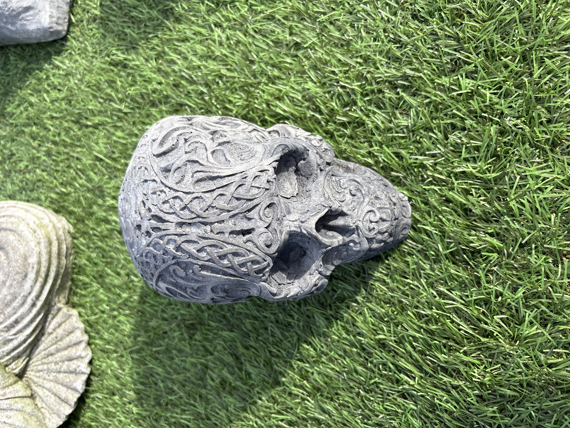 The image depicts a gray, stone skull ornament resting on a patch of artificial green grass. The skull is intricately carved with Celtic knot patterns. A portion of a gray, shell-shaped stone object is visible on the left side of the frame, also resting on the artificial grass. The scene appears to be outdoors in daylight. Although the specific location in Chirk, United Kingdom cannot be identified from the visual data, this is likely an outdoor decorative display.