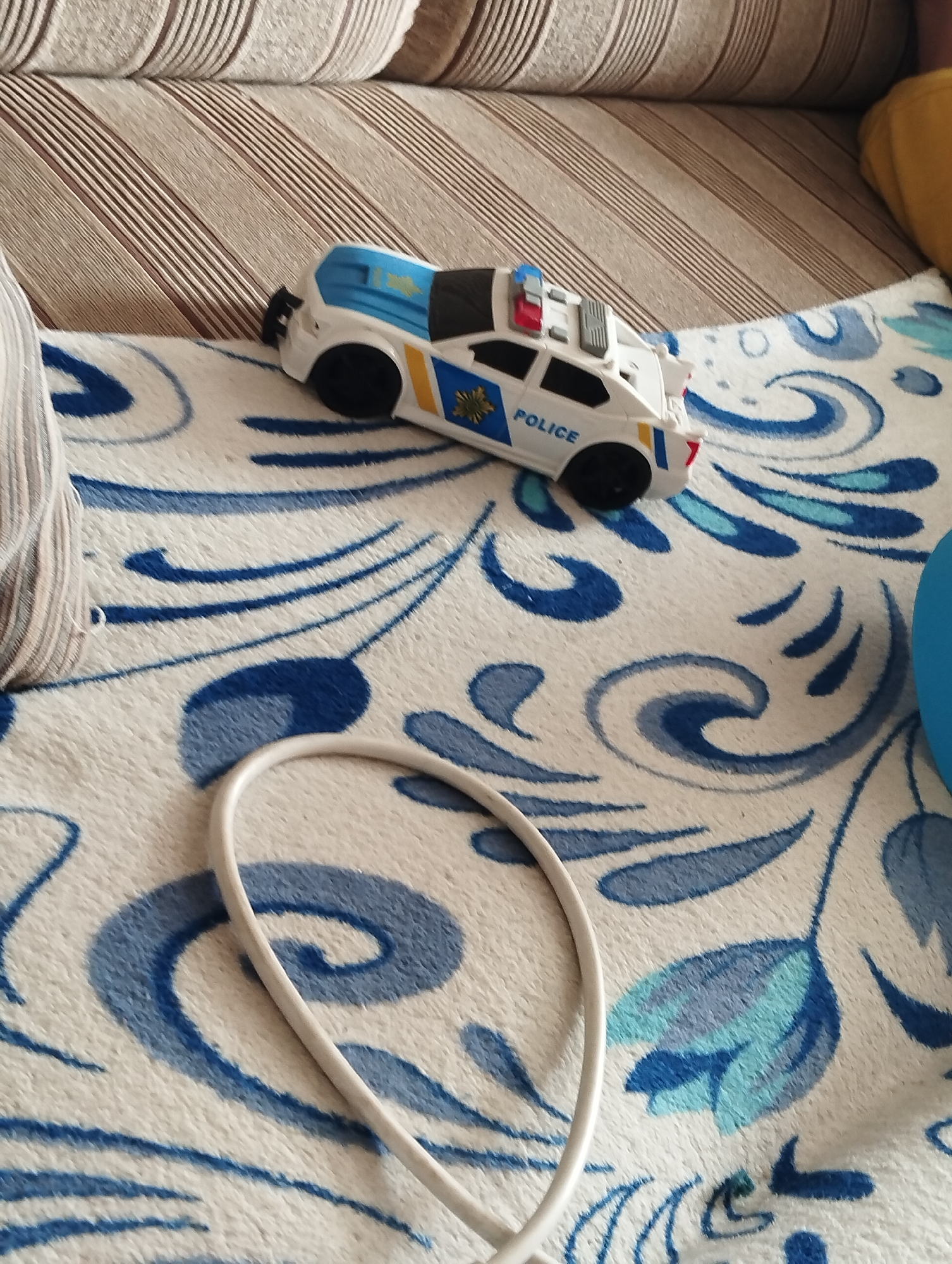 A white toy police car with blue and yellow accents sits on a white blanket with blue floral patterns. The car has "POLICE" written on its side in blue letters. The car is positioned on its side with its wheels facing upwards. The blanket is draped over a couch, and the scene appears to be indoors. A beige and brown striped couch is visible in the background, and a white cable lies on the blanket in the foreground. The lighting suggests daytime.
