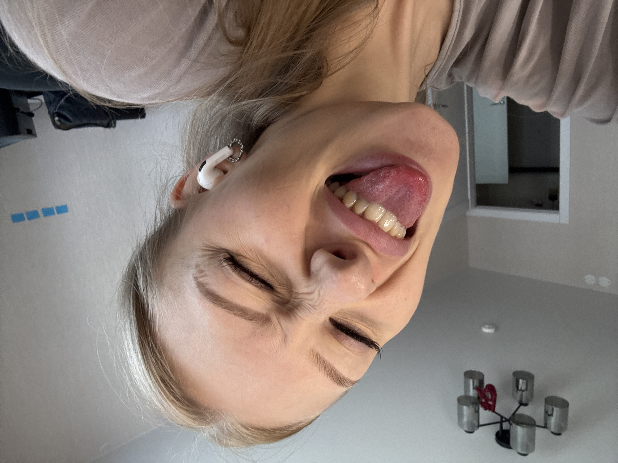 The image shows a close-up, selfie-style shot of a young woman with blonde hair and fair skin. She is sticking her tongue out and wincing, with her eyes squinted shut and wrinkles around them. She is wearing a light-colored top and white wireless earbuds in her right ear. A circular, sparkly earring is also visible in her right ear. The background is slightly out of focus but appears to be an indoor residential setting. Visible elements include a light-colored wall, a doorway with a white door, and a ceiling light fixture with multiple cylindrical lamps. There is also a dark object, possibly a bag, and a black box-like object in the background. The overall scene is consistent with a casual, personal moment captured at home. The location is not discernable from the image, and therefore cannot be associated with the city of Perm, Russia.