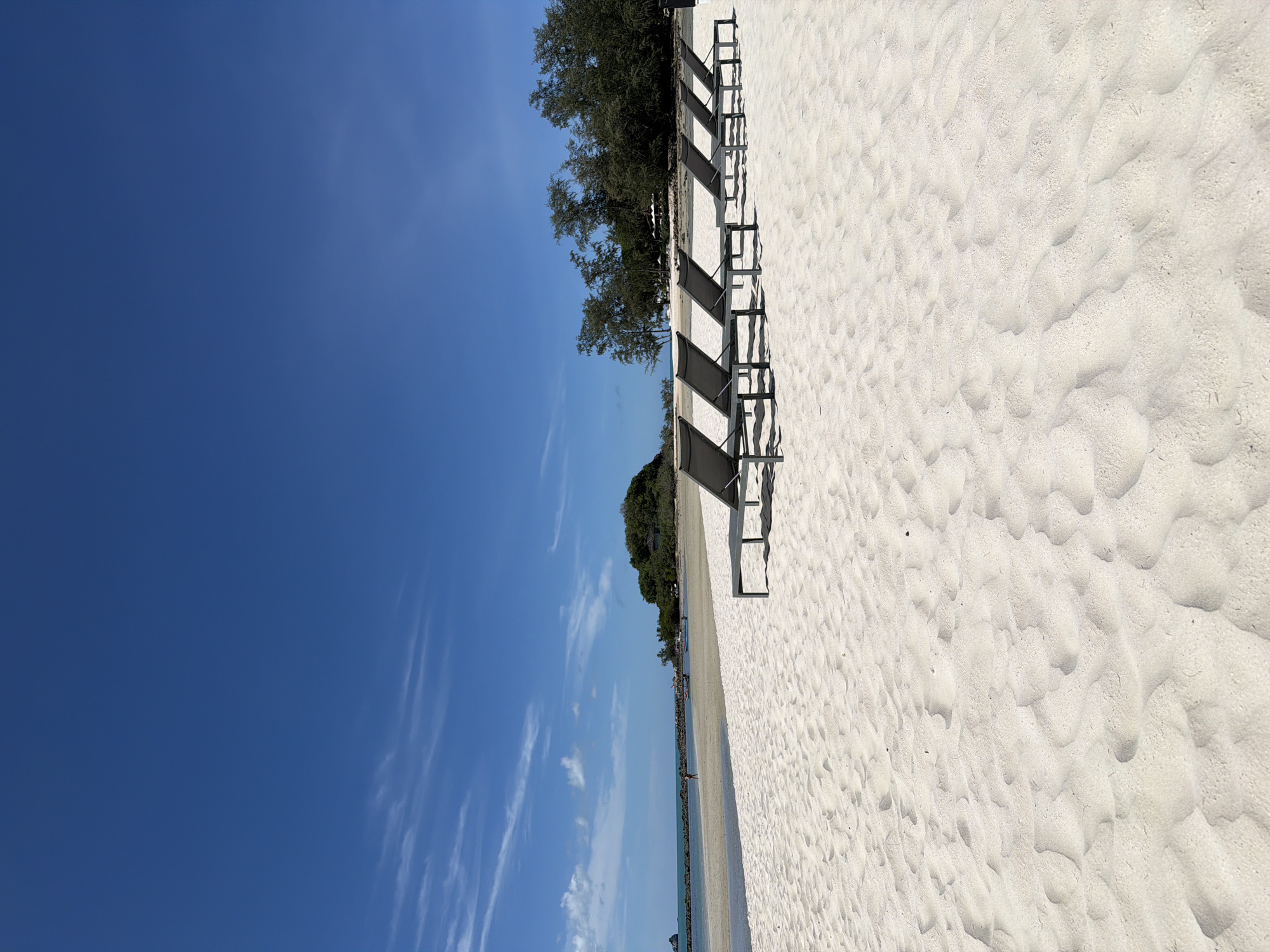 This is a vertical, eye-level, full shot of a serene beach in Baan Plai Laem, Thailand, on a bright, clear day with a deep blue sky and scattered white clouds. The foreground is dominated by pristine white sand with subtle ripples from the wind, creating a soft, textured surface. A row of eight dark grey lounge chairs is neatly arranged on the sand, positioned to face the ocean.
In the midground, a calm body of turquoise water meets the shore, with a small sandbar or shallow area creating a natural divide. A single person in a red outfit is walking along the water's edge, facing away from the camera. Further in the distance, a small, tree-covered island with a single building on it is visible. A few small boats are anchored near the island. The background features a dense line of trees, suggesting a tropical landscape beyond the beach. The scene conveys a sense of tranquility and tropical relaxation.