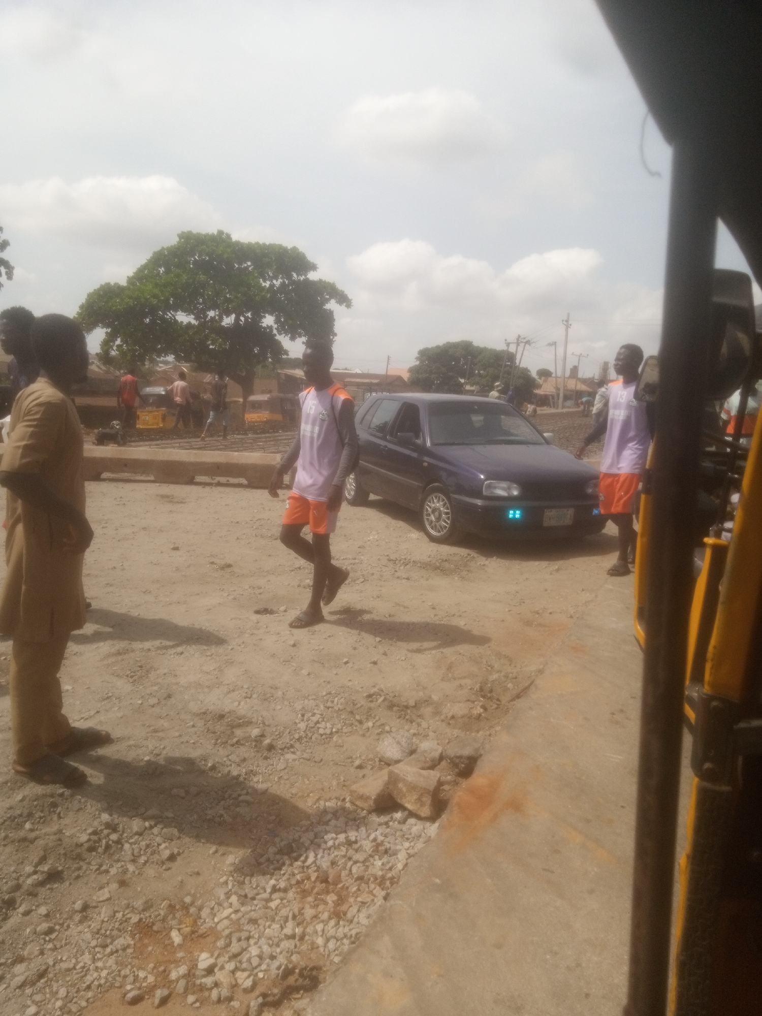 In Nassarawa, Nigeria, under a bright, cloudy sky, a scene unfolds on a dirt and gravel road. Three young men are the central figures. On the left, a man in a tan uniform stands with his back to the camera, facing away from the road. To his right, a man in a white and orange athletic shirt and orange shorts walks across the road, his gaze directed forward. Further to the right, a third man, also in a white and orange athletic shirt and orange shorts, stands near a dark blue Volkswagen Golf car. The car has its headlights on, and blue LEDs are visible on the front bumper. In the background, a large tree with green foliage stands tall, and behind it, a few buildings and utility poles are visible. Several yellow tricycles are parked or moving in the distance, suggesting a busy area. The ground is uneven, with patches of sand, gravel, and some scattered rocks. A section of a yellow vehicle, possibly part of a motorcycle or a tricycle, is visible in the foreground on the right side of the image. The overall impression is a snapshot of everyday life in a Nigerian city.