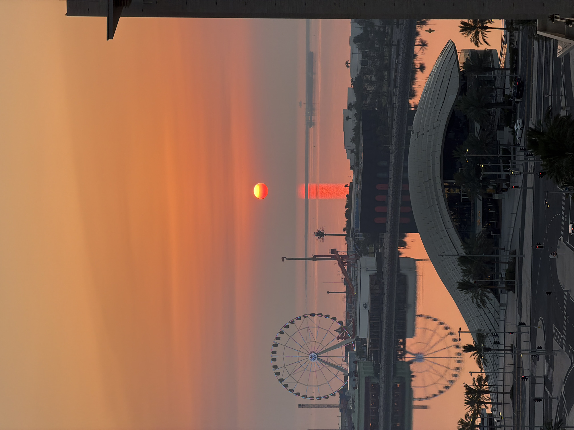 An aerial view captures a vibrant sunset over a coastal city. The sky is a gradient of warm orange and pink hues, with the sun, a glowing red orb, positioned slightly to the left of the frame, casting a fiery reflection on the calm water. In the foreground, a modern architectural building with a curved roof is visible, surrounded by palm trees and a street intersection with traffic signals. Across the water, a large Ferris wheel with illuminated spokes stands prominently against the horizon, part of an amusement area with other structures and rides. Distant ships are faintly discernible on the water. The overall scene depicts a serene urban landscape bathed in the warm glow of twilight.