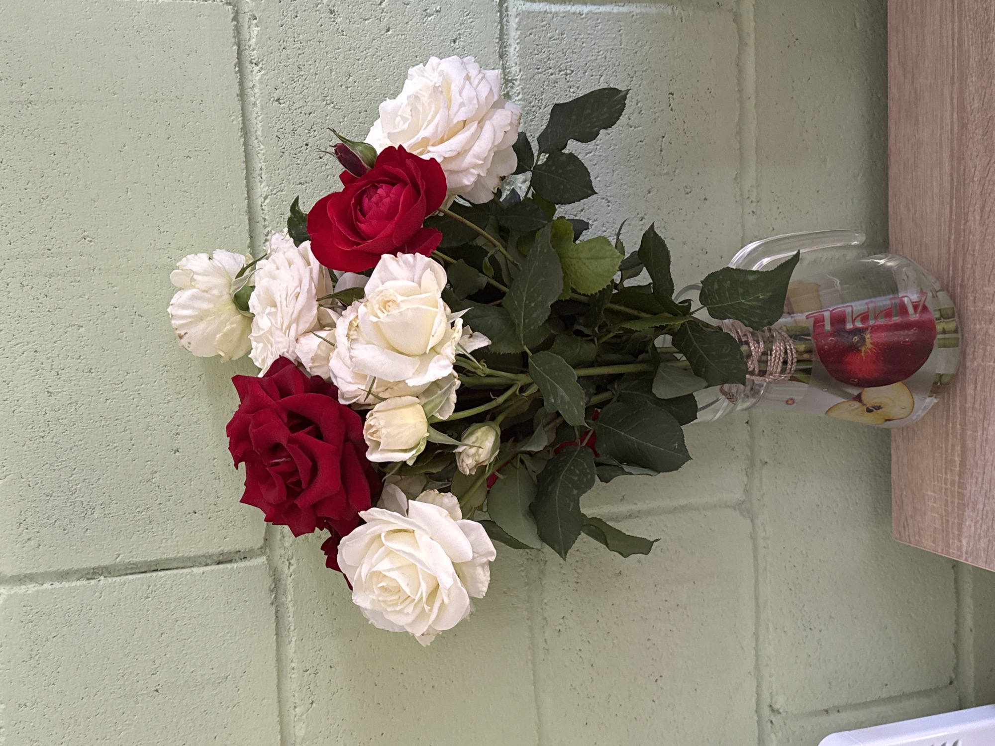 A bouquet of red and white roses is arranged in a clear glass vase decorated with an apple motif. The vase sits on a light-colored wooden surface. Behind the vase is a textured light green wall constructed from cinder blocks. The image does not depict any people or actions.