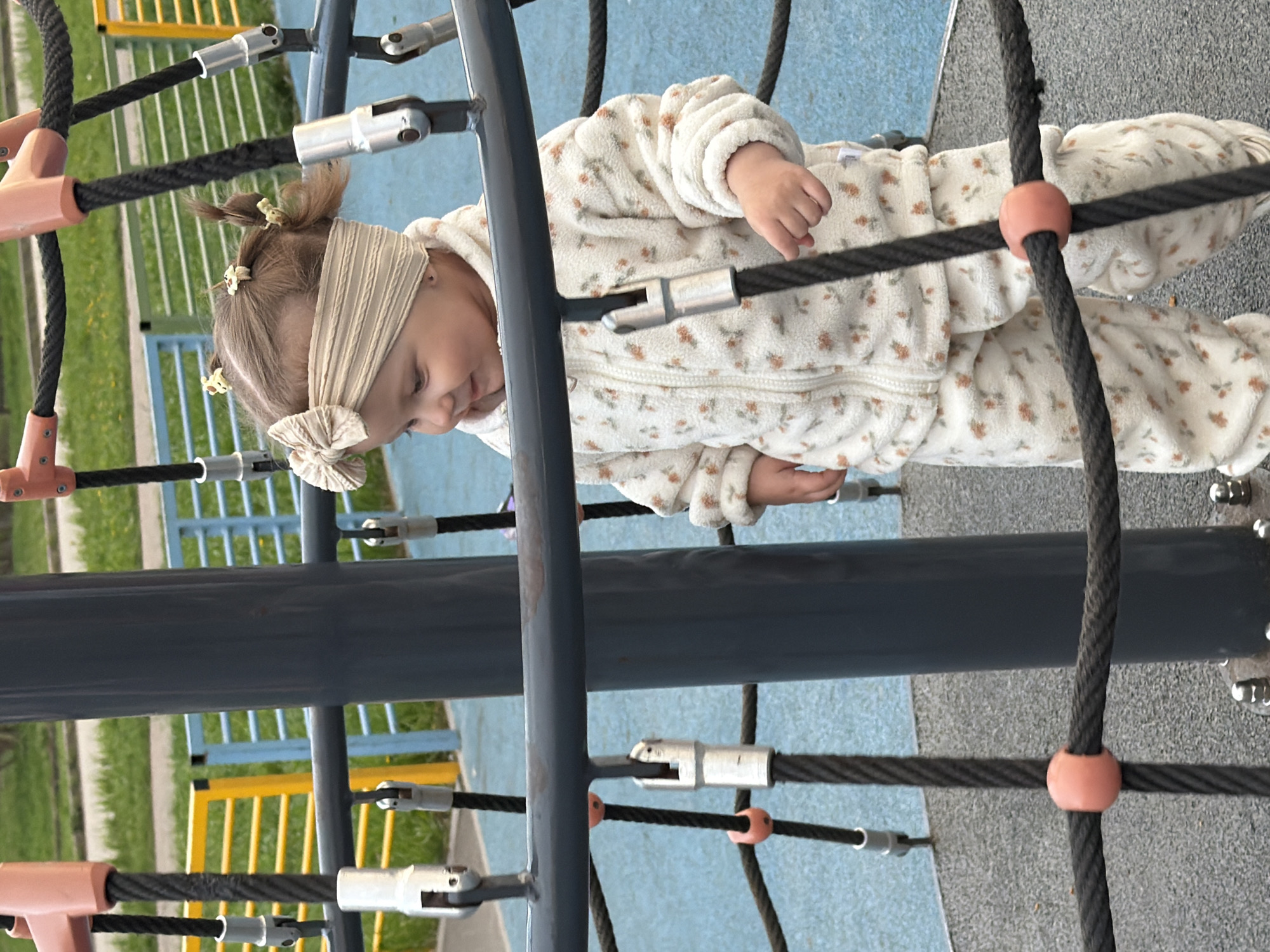 A young child in a floral-patterned jumpsuit and headband is on a rope-climbing structure. The location appears to be a playground. In the background, there is grass, fencing, and blue and yellow playground equipment. There is no information to confirm the location is in Reshetnikovo, Russia.