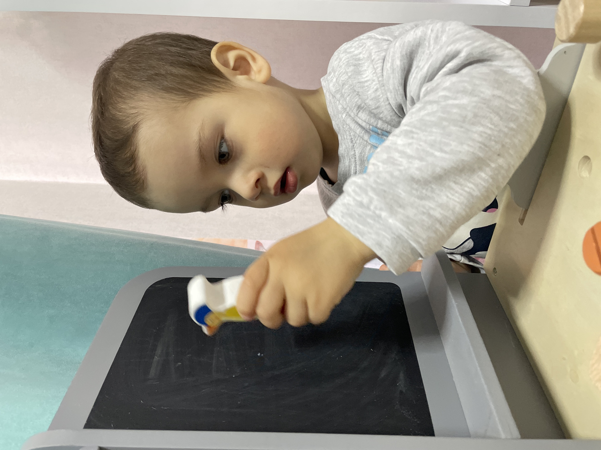 A young child, likely between 2-4 years old, stands in front of a chalkboard. He holds a white, yellow, and blue object in his right hand, and appears to be drawing or erasing on the board. The child has fair skin, light brown hair, and wears a gray long-sleeved shirt. The chalkboard is set in a light gray frame. Part of a wooden structure with orange circles is visible on the right side. The background includes a light blue surface and hints of a white shelf or wall. There's no information in the image to determine if the photo was taken in Izhevsk, Russia.