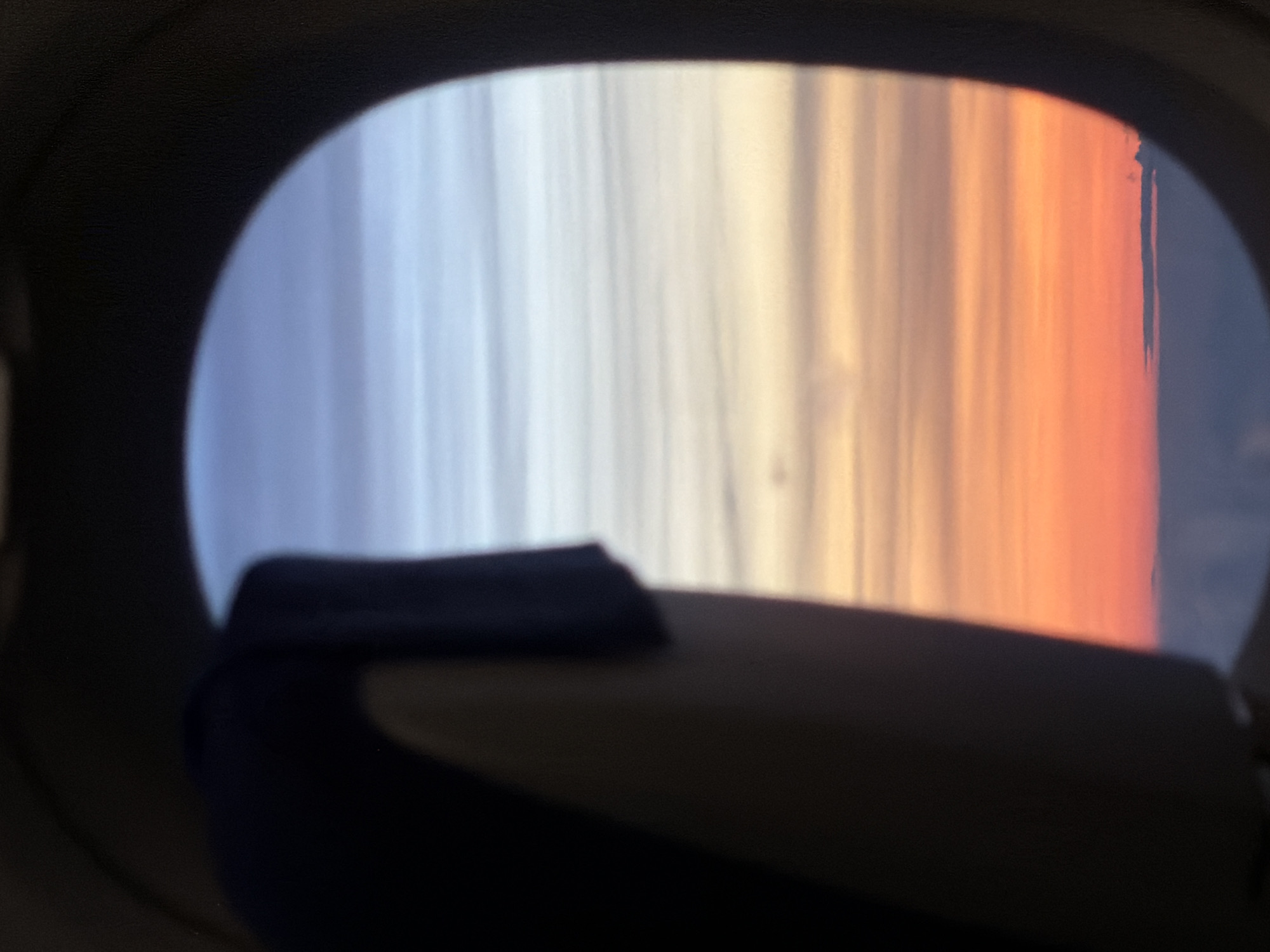 The image is taken from an airplane window, looking out at a sunset. The foreground is dominated by a dark shape, likely part of the airplane's interior, with a dark blue headrest visible. The window frames a view of the sky, which transitions from a bright orange and pink near the horizon to lighter shades of blue and white higher up. Thin, horizontal cloud formations are visible, creating layered stripes across the sky. The horizon line is low and appears to be above a layer of clouds. There are no discernible landmarks or features that would indicate a specific location within Nadergul, India, or any city. The scene is a typical view of a sunset from an airplane.