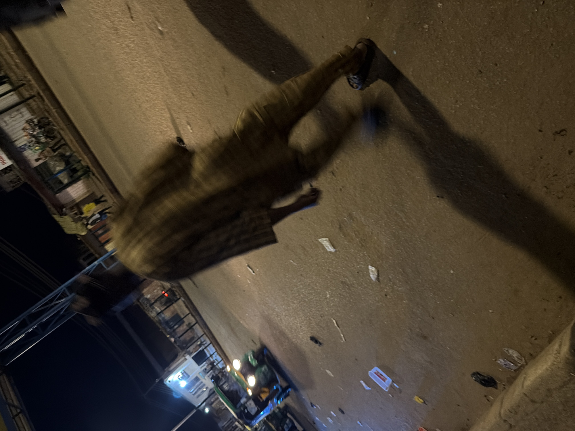 The image shows a man wearing a patterned shirt and trousers walking on a dirty street at night. There is a tuk-tuk vehicle with its headlights on, moving away from the viewer, on the left side of the image. The street is littered with various debris, including plastic bottles and paper. In the background, there are buildings with lights on, suggesting a commercial or residential area. The overall lighting suggests it is nighttime, possibly in an urban environment. The scene is consistent with a street in Bukuru, Nigeria, given the presence of a tuk-tuk, which is a common mode of transport in that region.