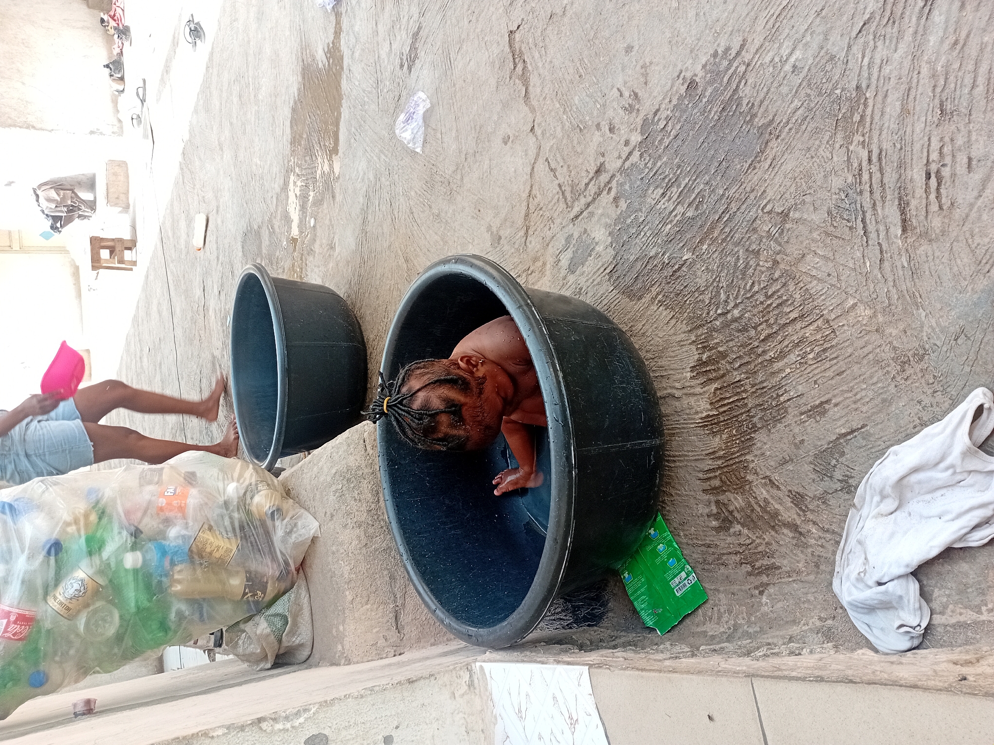 A young child sits in a large, black plastic tub with their head bowed down. The child appears to be bathing. The tub is positioned near a concrete wall and tile. An opened green packet is next to the tub. A second tub sits further away. Legs and feet are visible near a bag of waste bottles. The location is Unknown, Unknown.