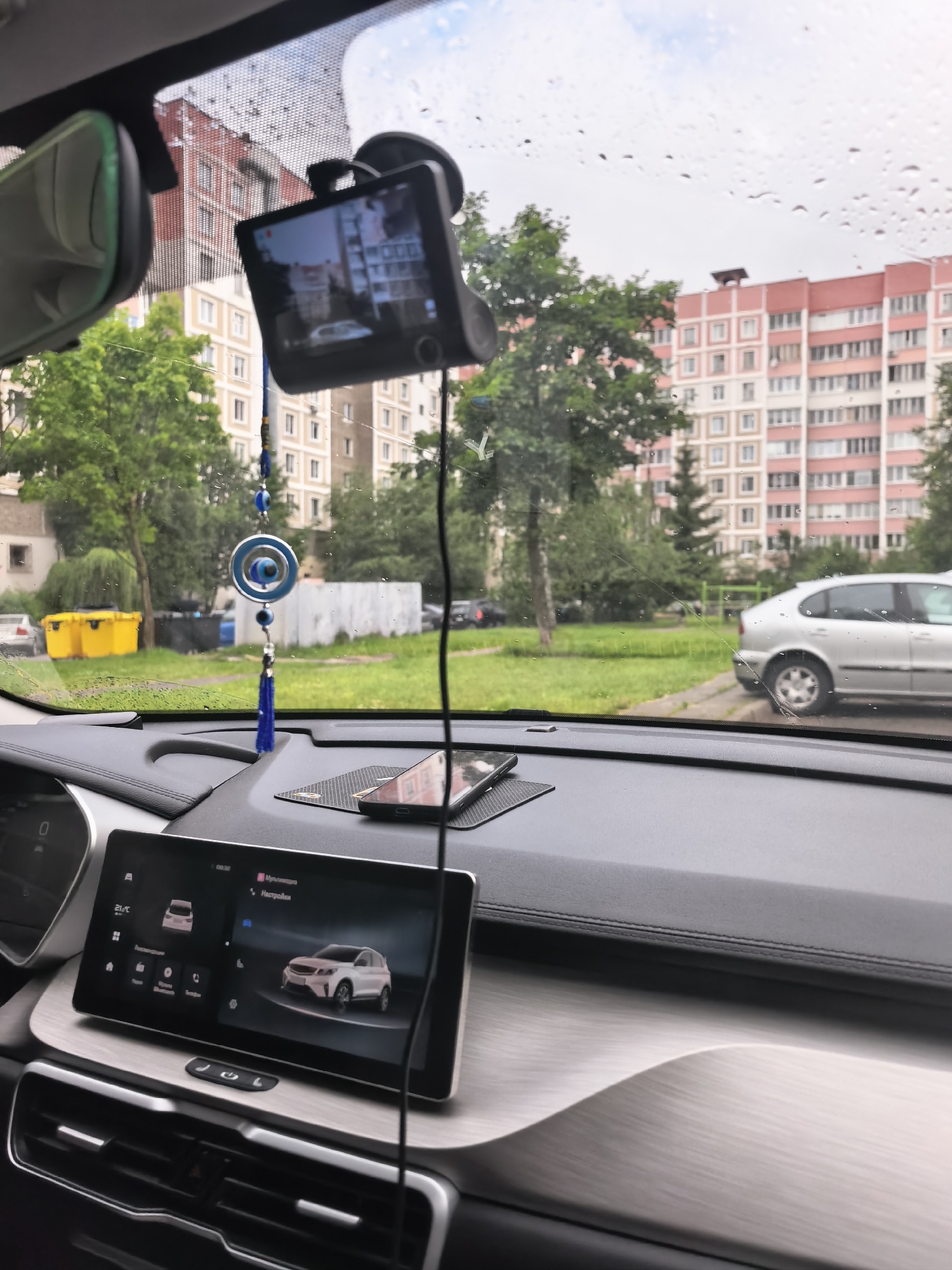 The image shows the interior of a car parked in an urban environment. Through the windshield, a row of apartment buildings and trees is visible. The sky is overcast with rain drops on the windshield. A dashcam is mounted on the windshield, and a smartphone is placed on the dashboard. A car's touchscreen infotainment system is visible in the foreground, displaying a car's interface. Outside the car, a silver sedan is parked on the side of the road, and two yellow trash bins are near a building. The scene appears to be in a residential area.