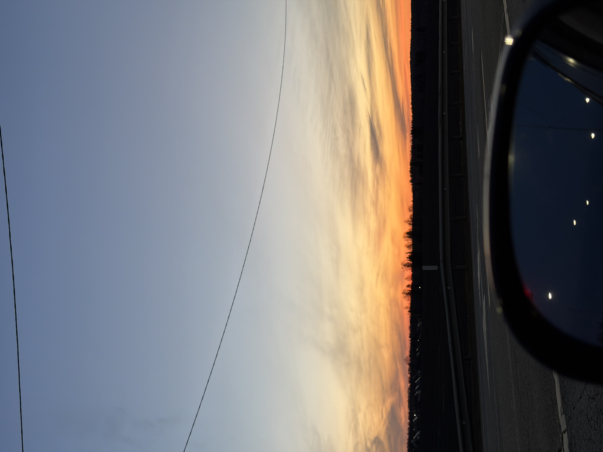 The image captures a roadside scene at twilight. On the right is a car's side mirror reflecting dim lights. Adjacent to the car, there's an asphalt road with visible markings and a barrier. Beyond the road, a line of trees is silhouetted against a vibrant sunset sky, transitioning from orange near the horizon to lighter hues higher up. On the left, the sky is a pale blue, with power lines visible. The specific location is Kuregovo, Russia, though no identifiable city elements or features are immediately apparent in the image.
