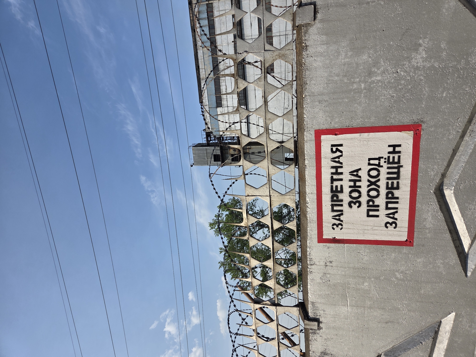 The image shows a concrete fence topped with hexagonal openings and barbed wire. A sign is attached to the fence with the inscription in Russian, "ЗАПРЕТНАЯ ЗОНА ПРОХОД ЗАПРЕЩЕН" (RESTRICTED AREA PASSAGE FORBIDDEN). Power lines are visible in the background, as well as a building. The sky is blue with some clouds. While it's impossible to confirm the specific location in Birobidzhan without further evidence, the presence of the sign in Russian suggests a location within a Russian-speaking area, potentially an industrial or restricted zone.