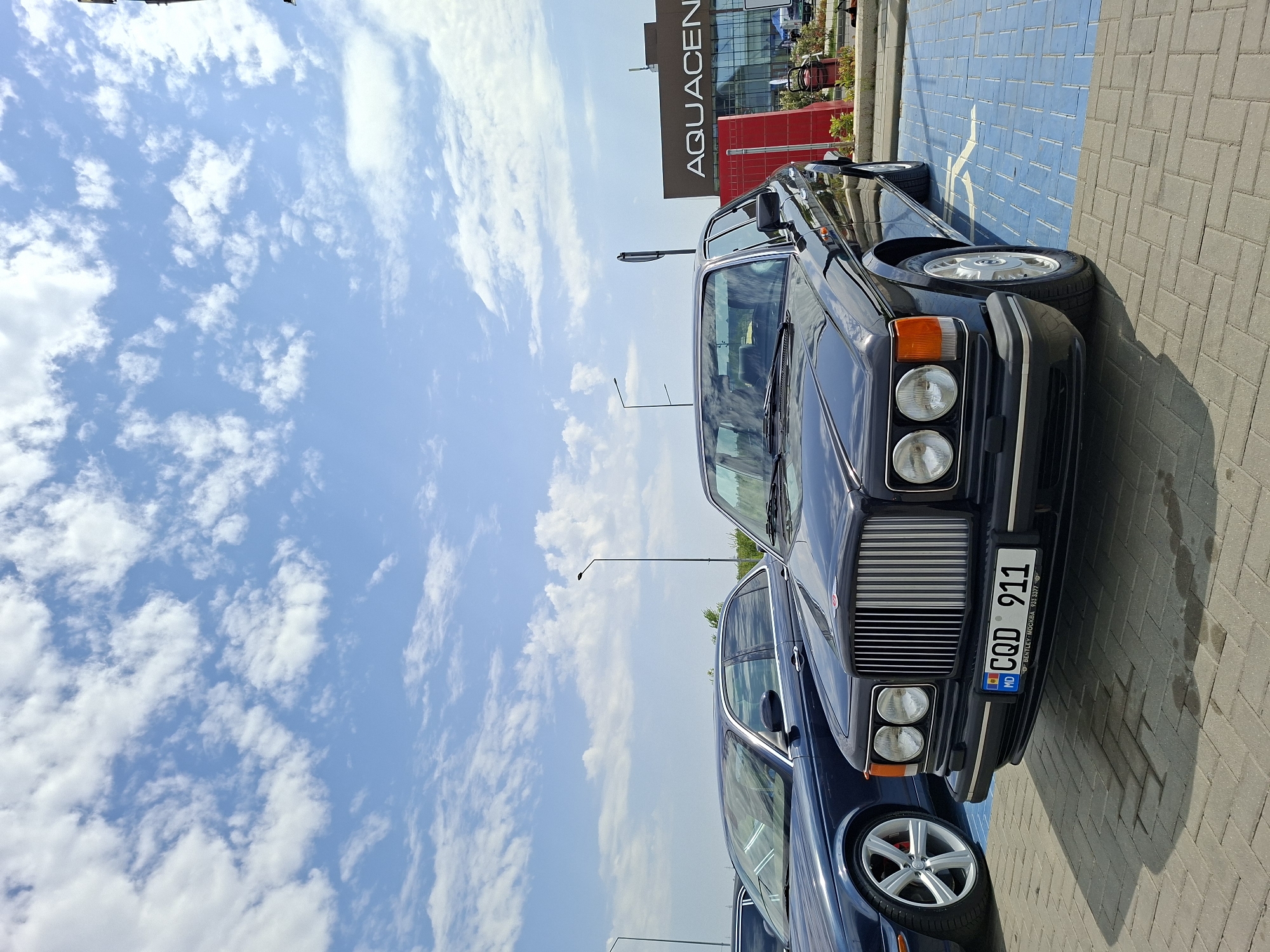The image depicts an outdoor scene, likely a parking lot, in Stăuceni, Moldova. The foreground features the front of a dark-colored Bentley, with its distinctive grille and headlights prominently displayed. The license plate reads "MD CQD 911". To the left of the Bentley, the front and wheel of a dark blue sedan are visible. The cars are parked on a patterned paved surface.
In the background, a modern building with the signage "AQUACENTER" is visible. The sky above is blue with scattered white clouds, indicating fair weather. Streetlights are present in the middle ground. No people are clearly visible in the frame. The overall impression is that of a car gathering or a showcase event.