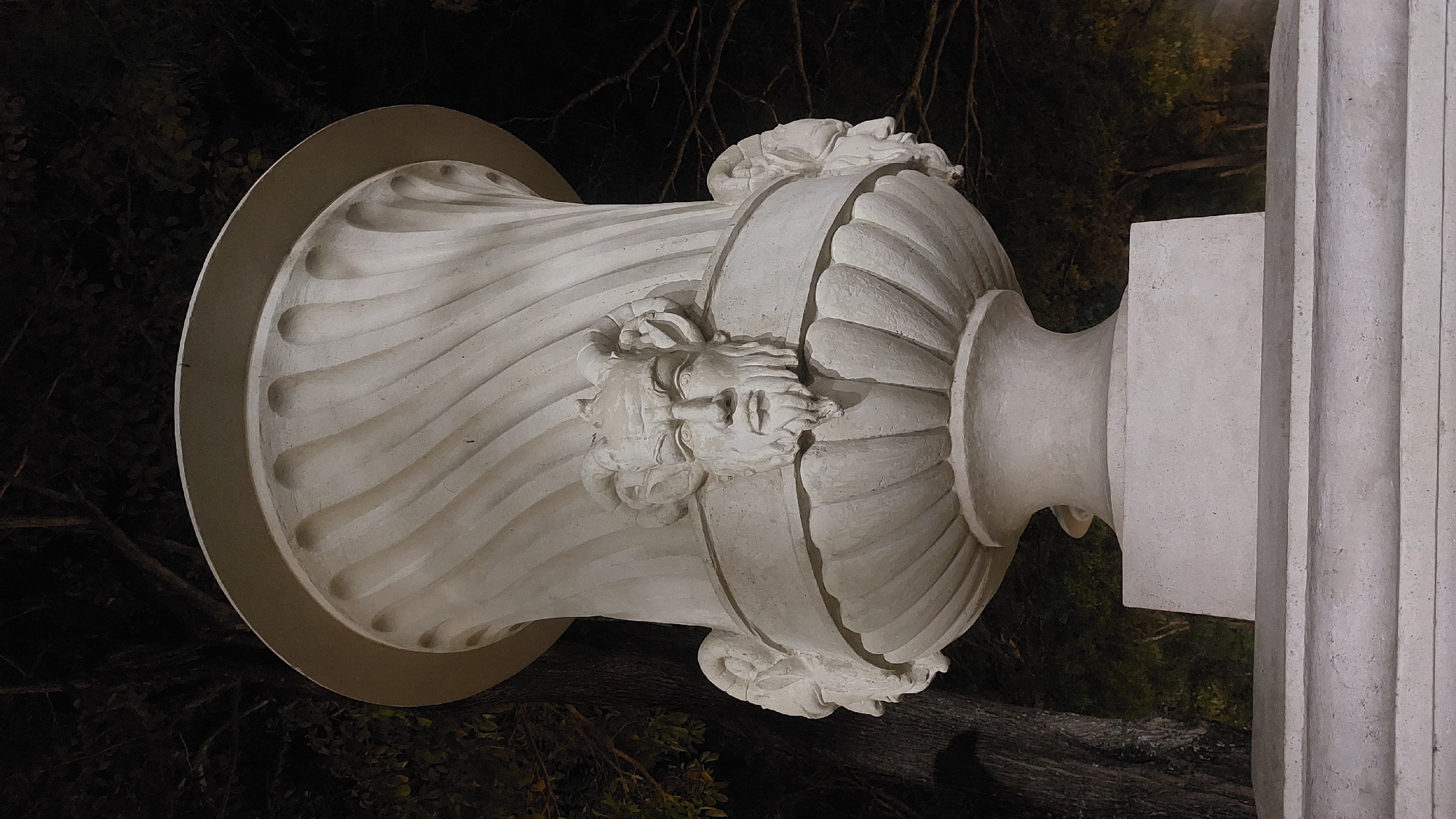 Here's a detailed description of the image:
The image shows a close-up of a white, ornate stone vase resting on a pedestal. The vase is decorated with carved faces and floral designs around its middle. The pedestal is simple, and its base has vertical grooves. The background is dark, with blurred trees and foliage suggesting the setting might be a garden or park. While I don't have the ability to pinpoint the exact location in Caracas, Venezuela based solely on this image, the style of the vase and the setting suggest it may be part of a public art installation or a feature within a historic park or garden.