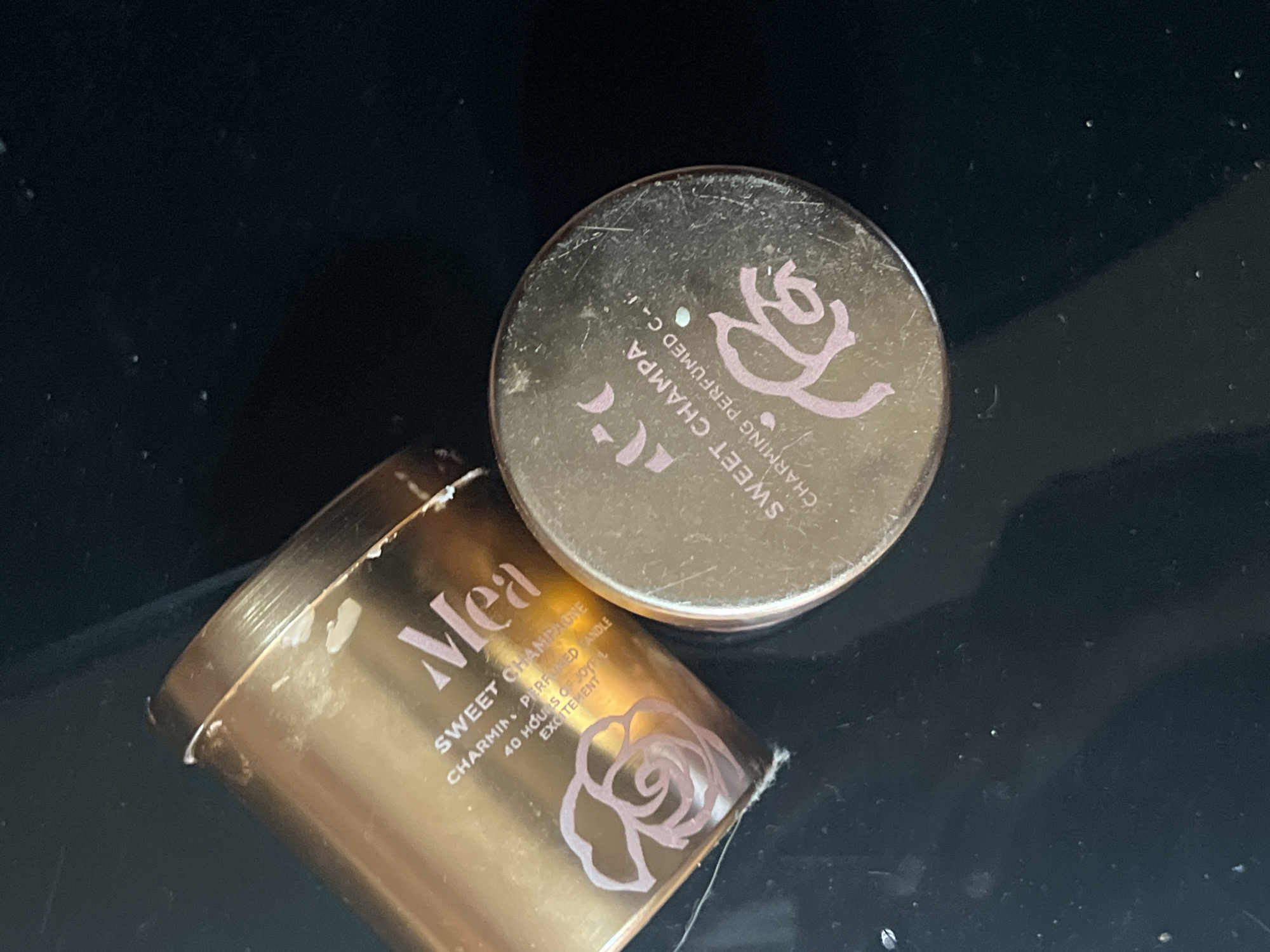 On a dark surface, possibly a table, are two gold-colored containers labeled "Mea" and "Sweet Champa," which appear to be related candle containers based on the text including "Charming Perfumed Candle" and "Charming Perfumed C-I." The label on the container reads "Mea SWEET CHAMPAGNE CHARMING PERFUMED CANDLE 40 HOURS OF JOYFUL EXCITEMENT". The container is open, with its circular lid situated nearby. The candle is likely from or inspired by the city of Boguchar, Russia, where one can buy and sell different products.