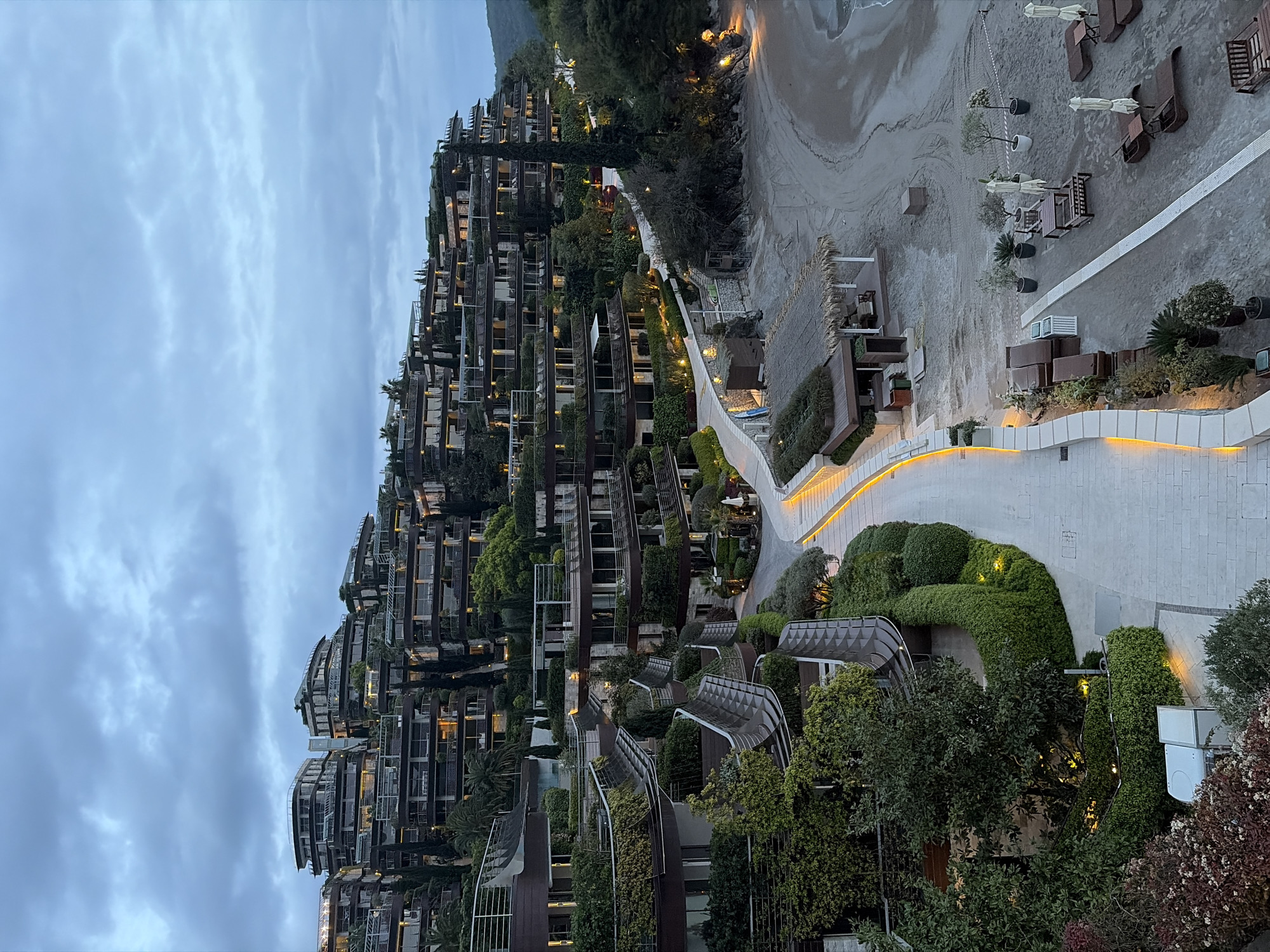 The image depicts a hillside hotel complex near the beach in the city of Budva, Montenegro. The terraced hotel structure is layered with greenery and multiple balconies. A paved pathway lined with illuminated landscaping separates the hotel complex from the sandy beach below. Beach furniture, including umbrellas and lounge chairs, are arranged on the beach. The sky is overcast and the scene appears to be at dusk or dawn.