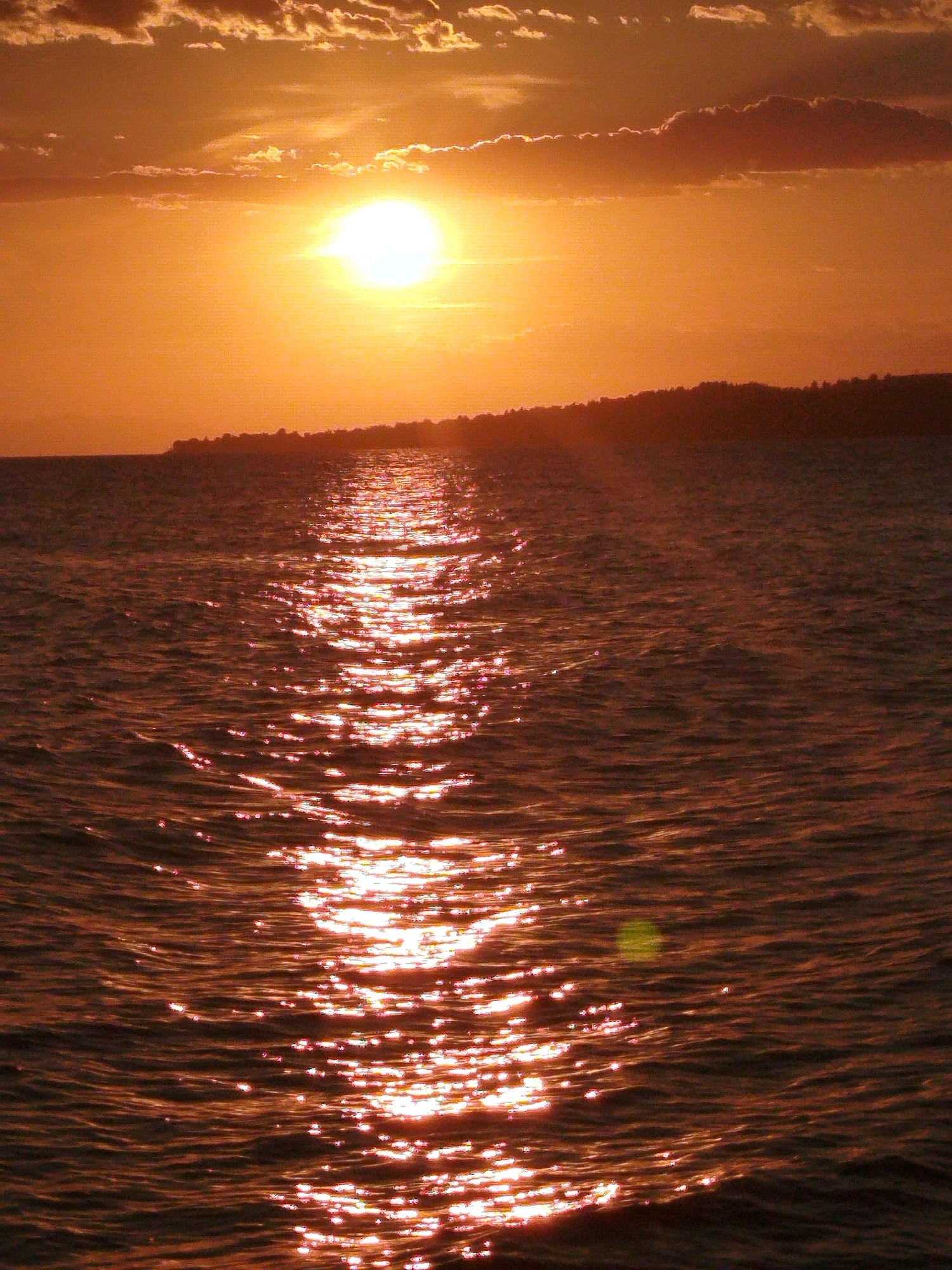 A sunset over the sea casts an orange glow across the scene. The sun, a bright orb, is positioned in the upper left quadrant of the image, partially obscured by clouds. A shimmering reflection of the sun stretches across the water's surface, creating a path of light that leads towards the viewer. The water is choppy, with small waves rippling across its expanse. In the background, a dark, silhouetted landmass lines the horizon, its details indistinct against the bright sky. The overall atmosphere is one of tranquility and natural beauty, typical of a coastal landscape in Sithonia Municipal Unit, Greece.