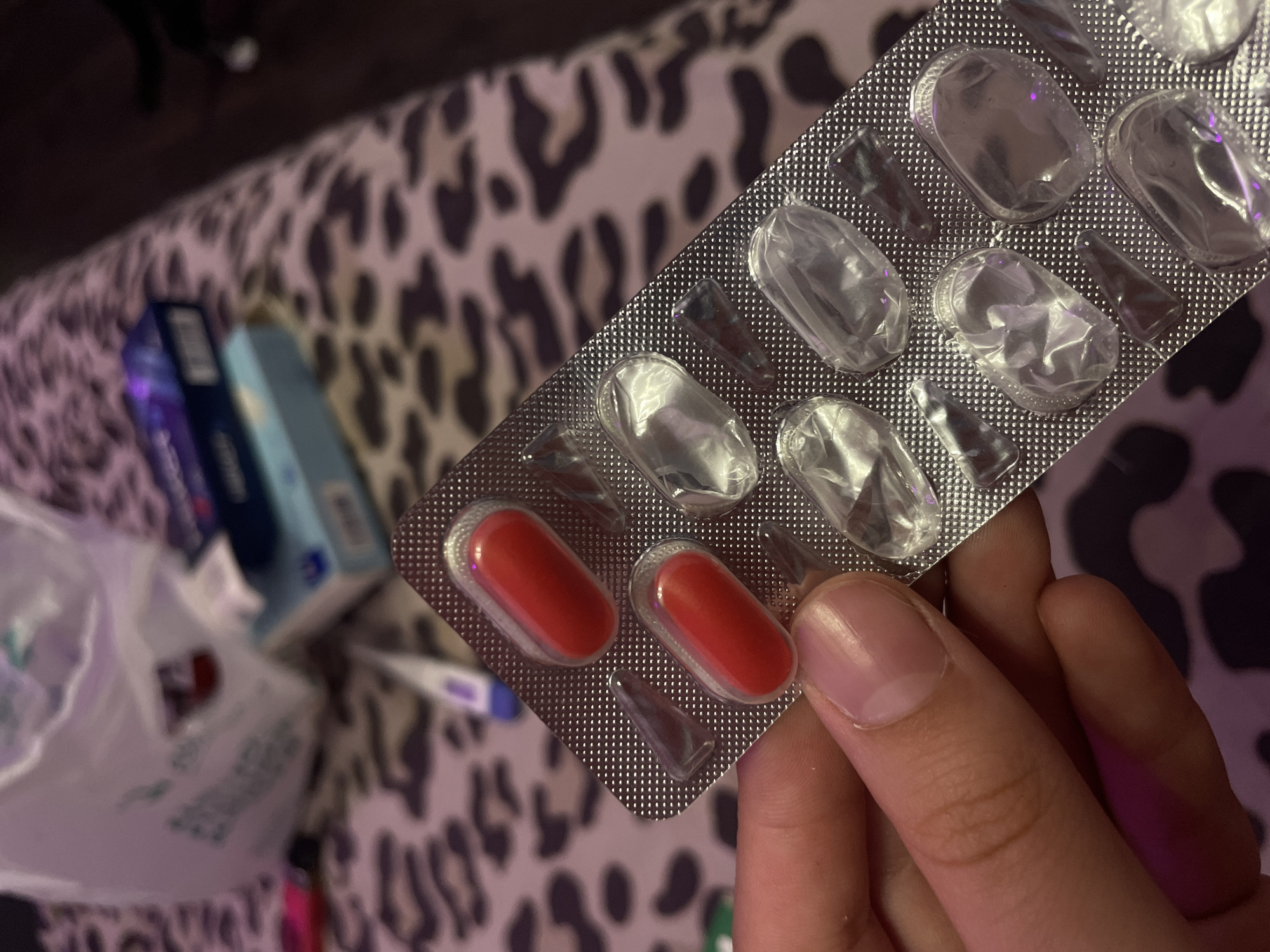 A hand holds a blister pack containing two red pills, with the remaining cavities empty. The blister pack is positioned in the foreground of the image. In the background, a leopard-print fabric is visible, serving as a surface. Behind the fabric, several items are blurred: a white plastic bag, a thermometer, and two boxes, one blue and one darker colored. The scene appears to be indoors, with artificial lighting. There is no information or indication of the location being Vaganovo, Russia.