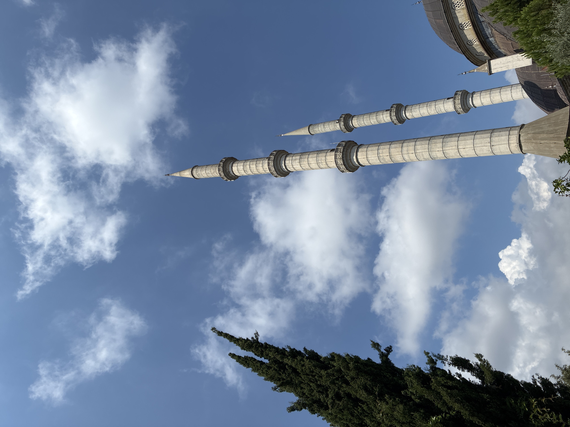 A low-angle, full shot captures the imposing minarets of a mosque against a bright blue sky dotted with white clouds. On the left, a tall, dark green conifer tree extends upward, its needles sharply defined. To the right, two slender, light-colored minarets rise prominently, featuring multiple balconies and pointed finials. Part of a domed mosque structure, clad in dark material and adorned with small arched windows, is visible behind the minarets. Green foliage from surrounding trees is also present in the lower right corner. The scene is devoid of people or any discernible activity. The architectural style and the presence of minarets strongly suggest this is a mosque, likely in Turkey. Given the context of Alanya, Turkey, this could be the Minarets of the Sultan Alaaddin Mosque or another mosque in the region.