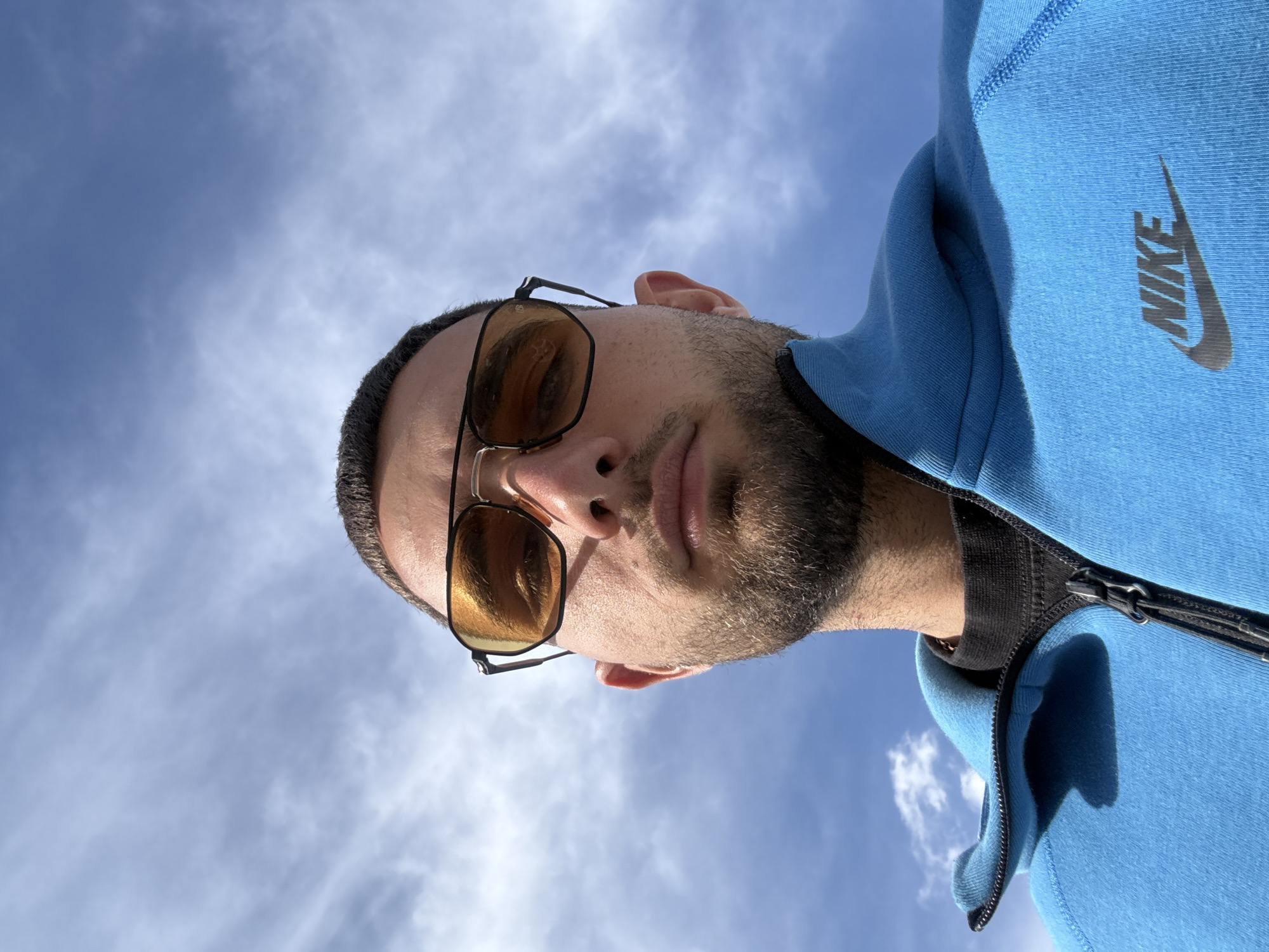 A man with short dark hair and a beard is looking down at the camera, wearing sunglasses with amber-tinted lenses. He has on a light blue Nike zip-up hoodie. The background is a bright blue sky with scattered white clouds. The location cannot be determined to be in Gabrovo, Bulgaria from this image alone.