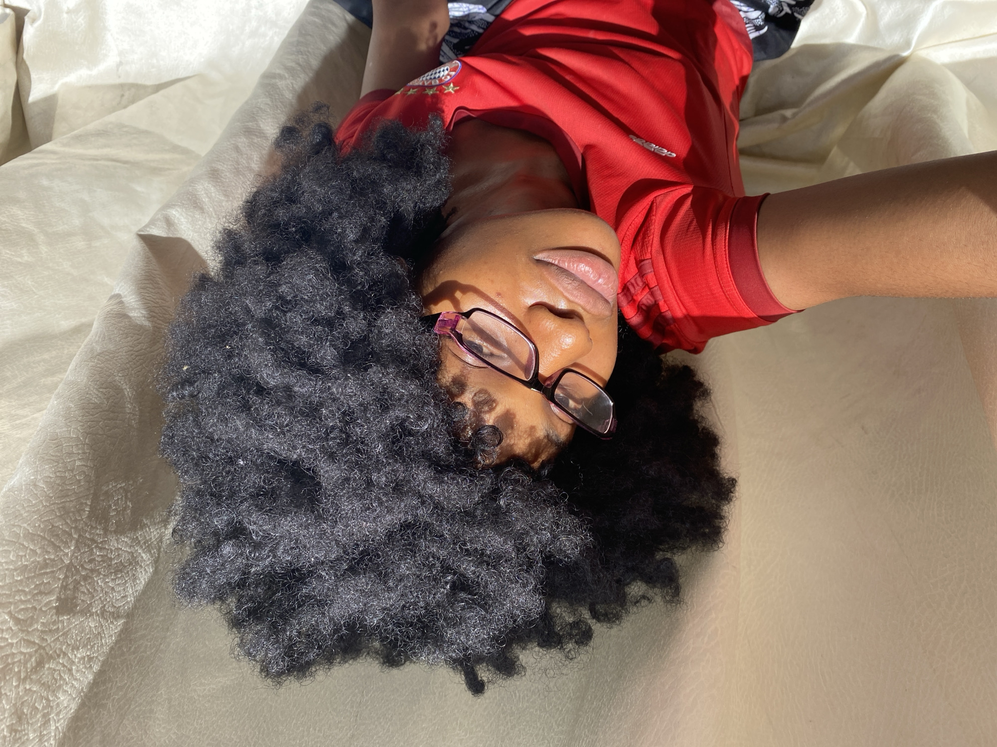 A woman is lying on a cream-colored surface, possibly a bed or a large piece of fabric. She is wearing a red shirt and glasses. Her hair is dark and voluminous. The lighting suggests an indoor environment with natural light. There is no evidence in the image to support the location being in Iranla, Nigeria.