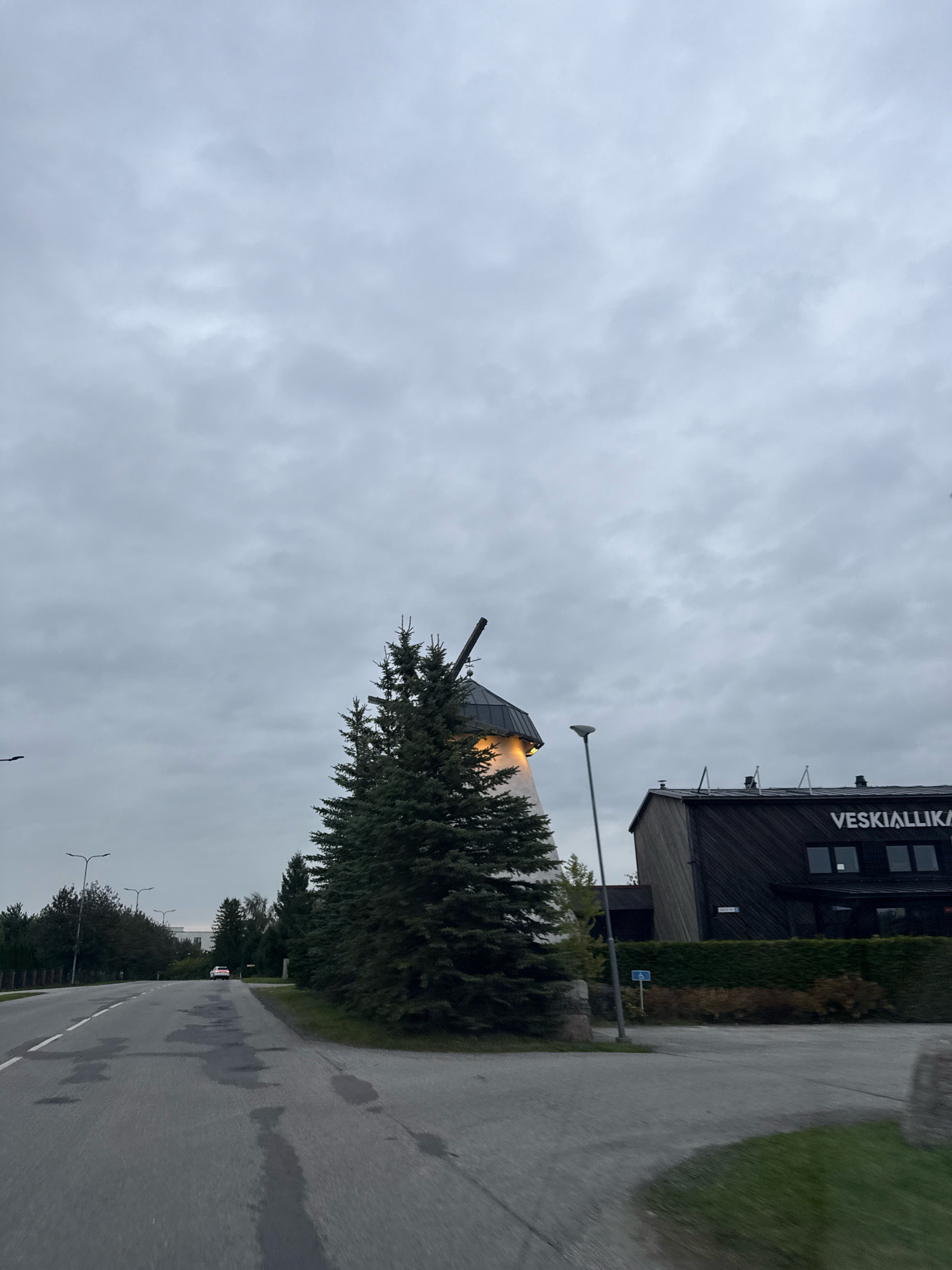 The image depicts an outdoor scene on an overcast day in Peetri alevik, Estonia. The foreground shows a wet asphalt road with white lane markings and several dark, irregular wet patches. To the left of the road, there are trees and streetlights. In the middle ground, a tall, dark evergreen tree stands prominently in front of a white-and-dark-colored building. Part of a windmill structure, with a black cap and illuminated windows, is visible behind the tree. To the right of the windmill, a dark, modern-looking building with the text "VESKIALLIKA" in white lettering is partially visible. The building has several windows. A streetlight is positioned in front of this building. The background consists of more trees and the cloudy sky. A white car is visible driving away on the road in the distance.