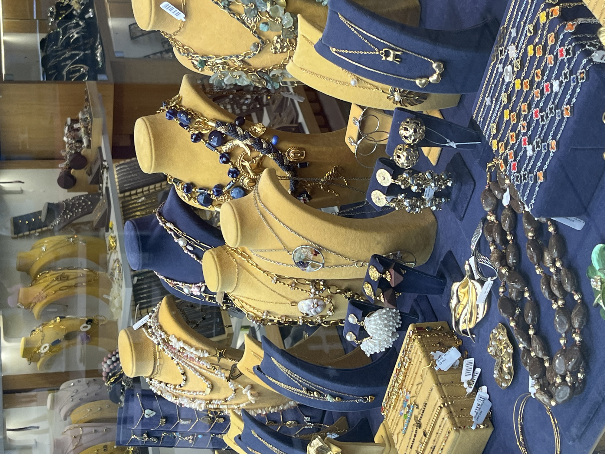 The image shows a storefront window display of jewelry, likely in Burgas, Bulgaria. The jewelry is arranged on yellow and navy blue display stands. Necklaces, earrings, and bracelets are visible, with a mix of gold, silver, and beaded designs. Some items have price tags attached. The overall impression is that of a retail environment selling fashionable accessories. The lighting suggests it is daytime.
