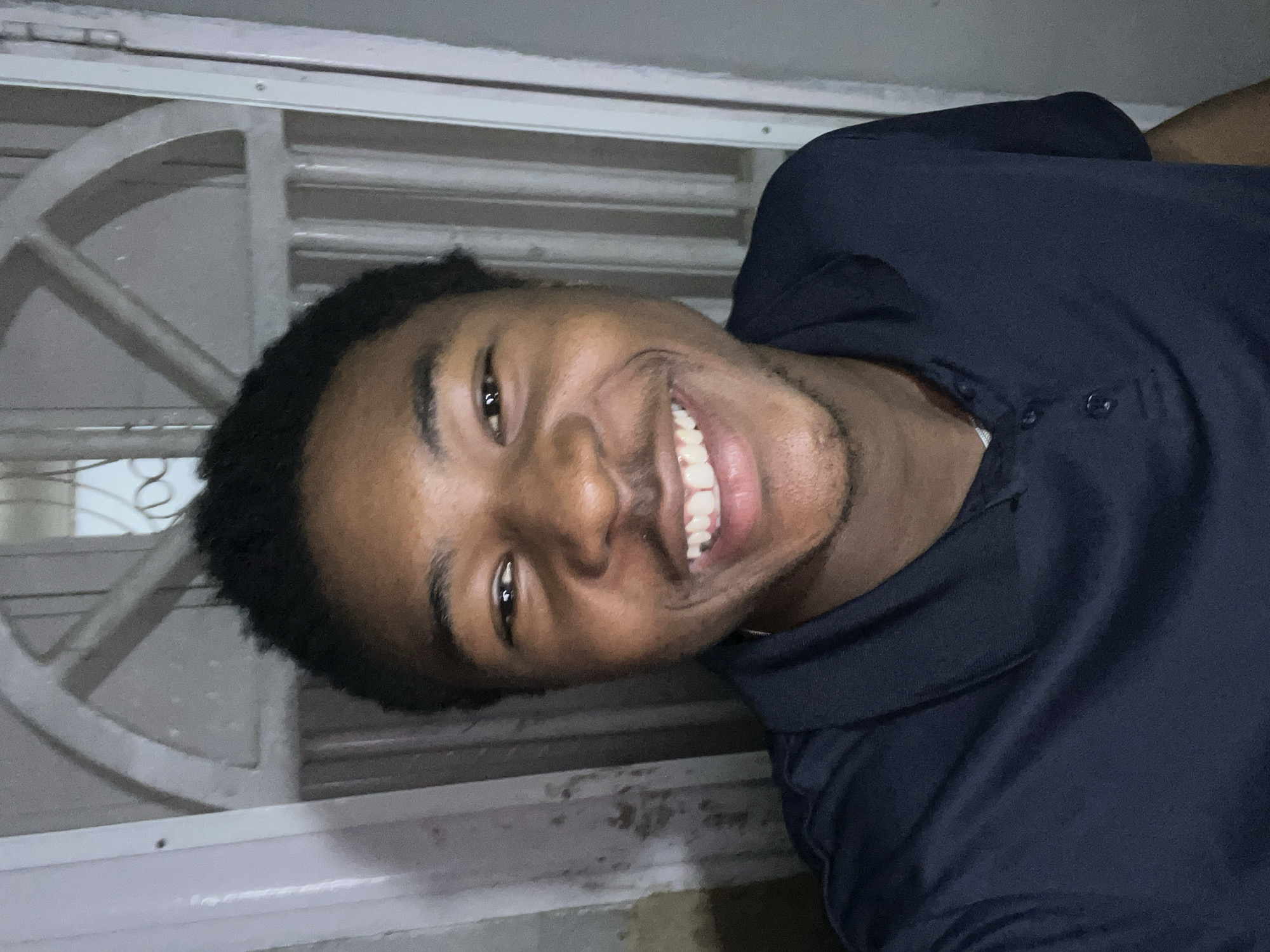 A young man smiles directly at the camera. He has dark hair and brown skin, and wears a dark blue polo shirt. Behind him is a window with white bars. The location is possibly in Jos, Nigeria.