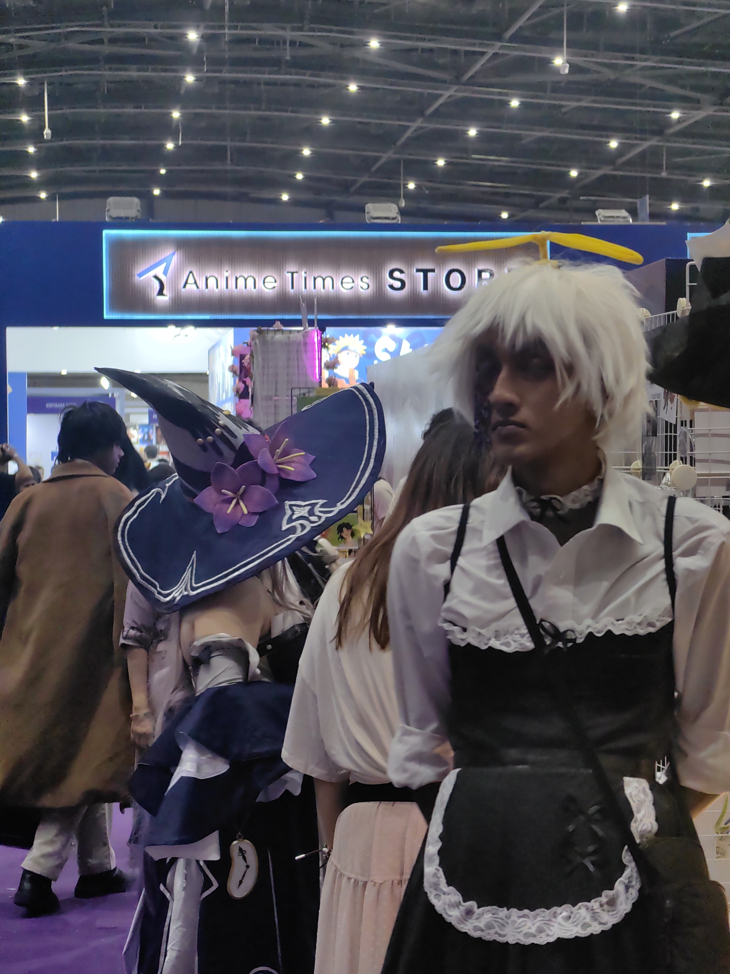 This is a medium shot of a group of people at what appears to be an anime or comic convention, likely indoors. In the foreground, a person with bright white, spiky hair and a yellow propeller hat is looking off to the right, wearing a white shirt with a ruffled collar and a black apron-like garment. To the left of this person, another individual is dressed in a dark blue witch's hat adorned with purple flowers and a dark blue and white outfit. Behind them, another person in a brown coat is visible. A sign in the background reads "Anime Times STOP." The lighting suggests overhead fluorescent lights, and the environment appears to be a large, open exhibition space with other attendees and displays in the background. The purple carpet underfoot is a notable detail of the floor covering. There is no specific indication of the city of Mumbai, India.