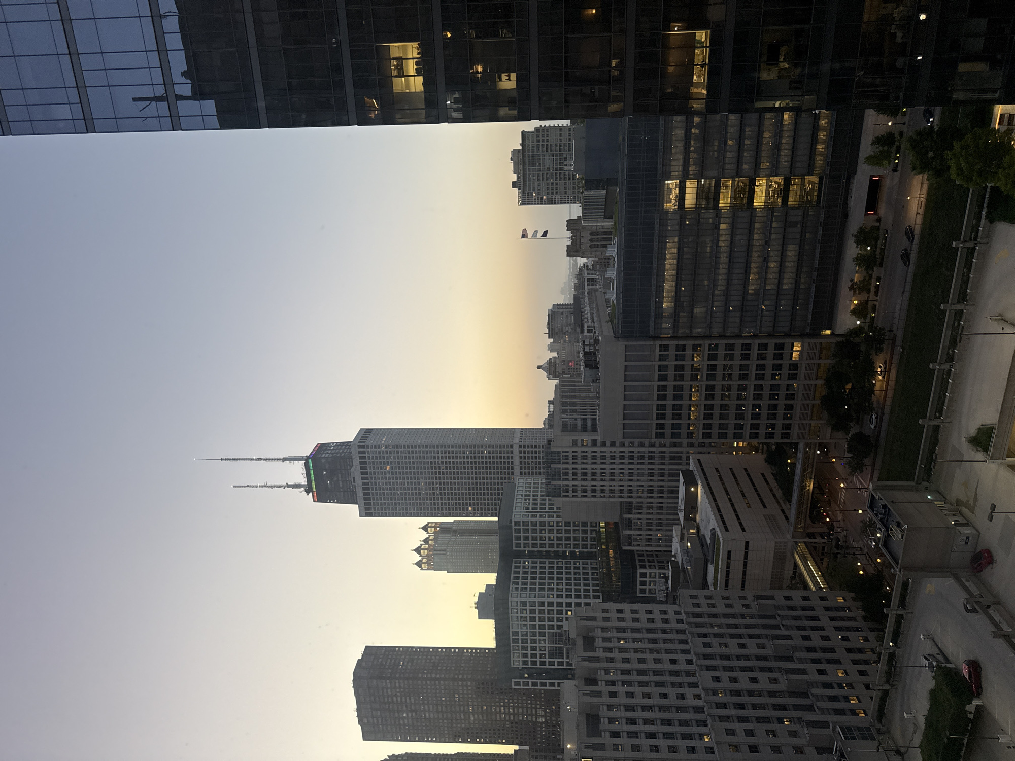 This image depicts a cityscape of Chicago, Illinois, USA, at dusk or dawn, with a clear sky transitioning from pale yellow to light blue. The foreground is dominated by the right side of a modern, dark-glass skyscraper, its numerous windows reflecting the sky and some internal lights.
In the mid-ground, a collection of buildings forms the urban landscape. The most prominent is the John Hancock Center (now 875 North Michigan Avenue), a tall skyscraper with two distinctive antenna masts. Its upper section is illuminated with a band of colored lights, possibly indicating a Pride flag or other celebration. To the left of the Hancock Center are other high-rise buildings of varying architectural styles, some with illuminated windows suggesting occupancy. A cluster of shorter, lighter-colored buildings with distinctive arched elements is visible behind and to the left of the Hancock Center.
Further into the background, more buildings are visible, some appearing as faint silhouettes against the horizon. Two flags are flying on a building in the mid-background, one appearing to be the American flag.
The lower portion of the image shows a multi-level parking structure with cars visible, and a street with lampposts and trees lining its edges. Some of the buildings in the mid-ground also show illuminated office spaces. The overall impression is a view of a dense urban environment during a period of low light.