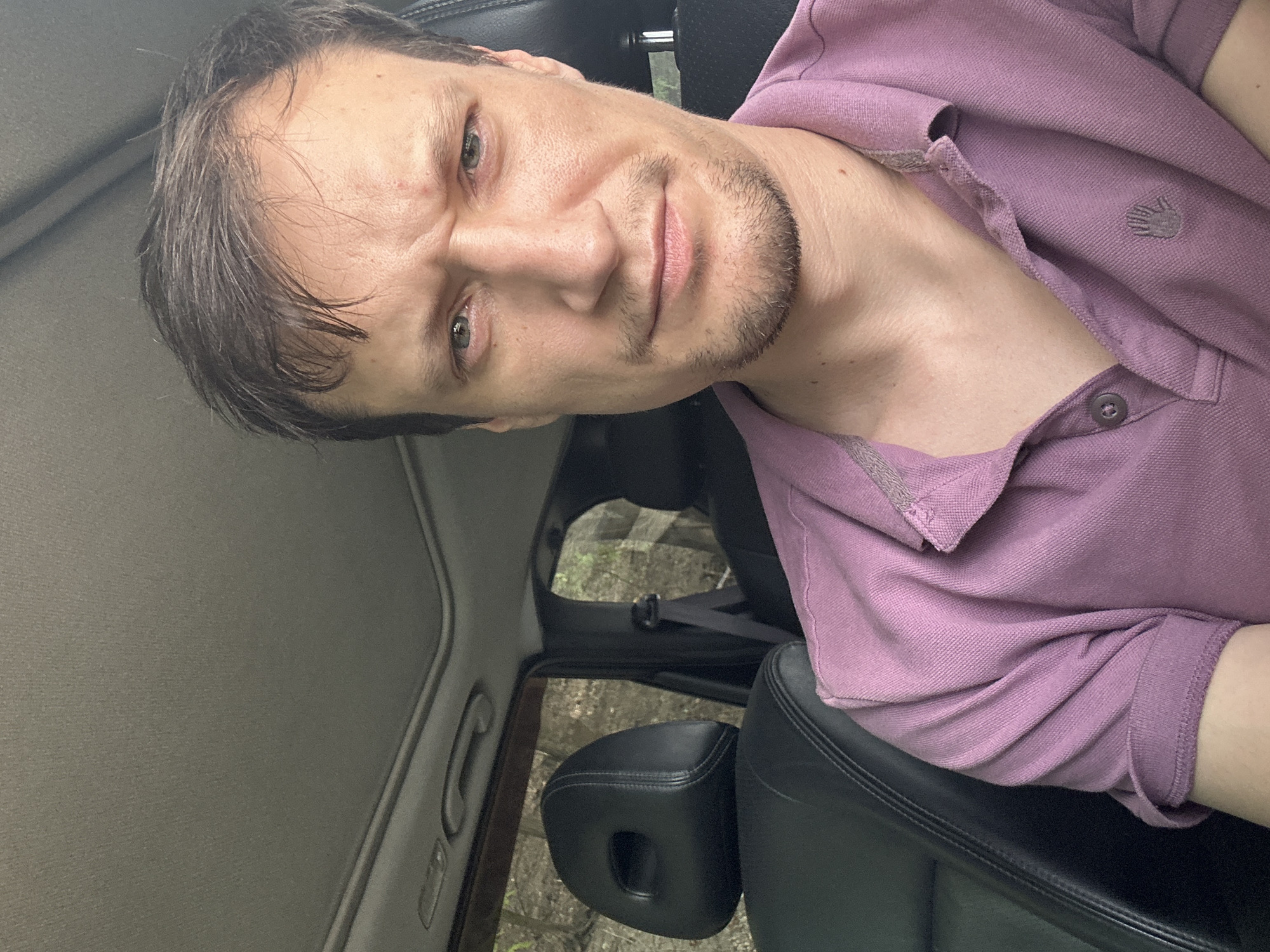 A close-up, eye-level, selfie-style shot shows a man sitting in the driver's seat of a vehicle, with his face filling the majority of the frame. The man, with light skin and short, dark brown hair, is looking directly at the camera with a slightly furrowed brow and a neutral expression. He is wearing a purple collared shirt. The interior of the car is visible, including a black leather headrest of a passenger seat behind him and the car's sunroof above. The background, seen through the car windows, appears to be a natural setting with trees and foliage, suggesting the car is parked or driving through a wooded area. The image is taken in Makiv, Ukraine.