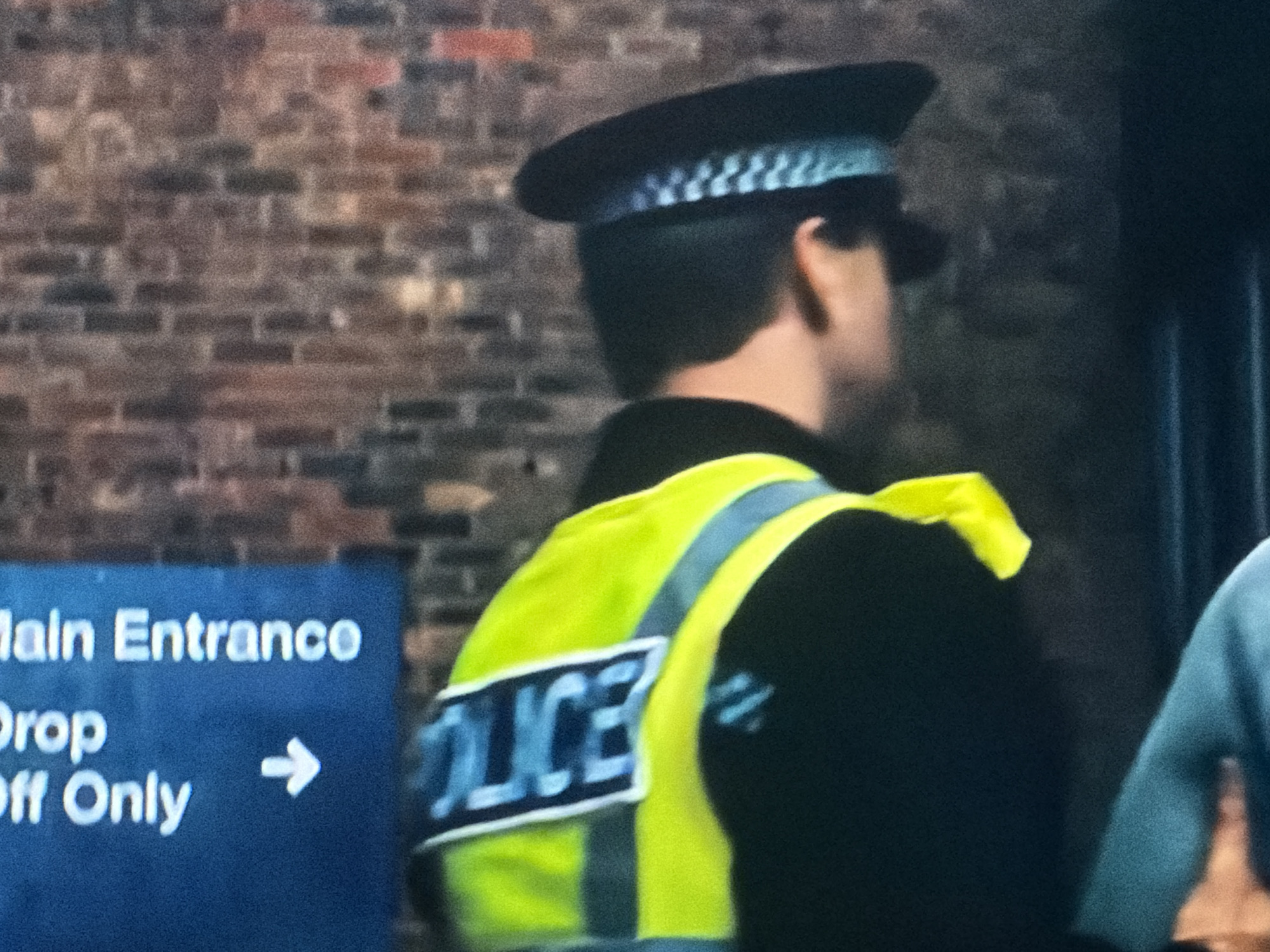 A police officer wearing a yellow high-visibility vest with "POLICE" printed on the back stands near a sign that reads "Main Entrance" and "Drop Off Only" with an arrow pointing right. The officer is wearing a police hat with a checkered band. The background includes a brick wall.