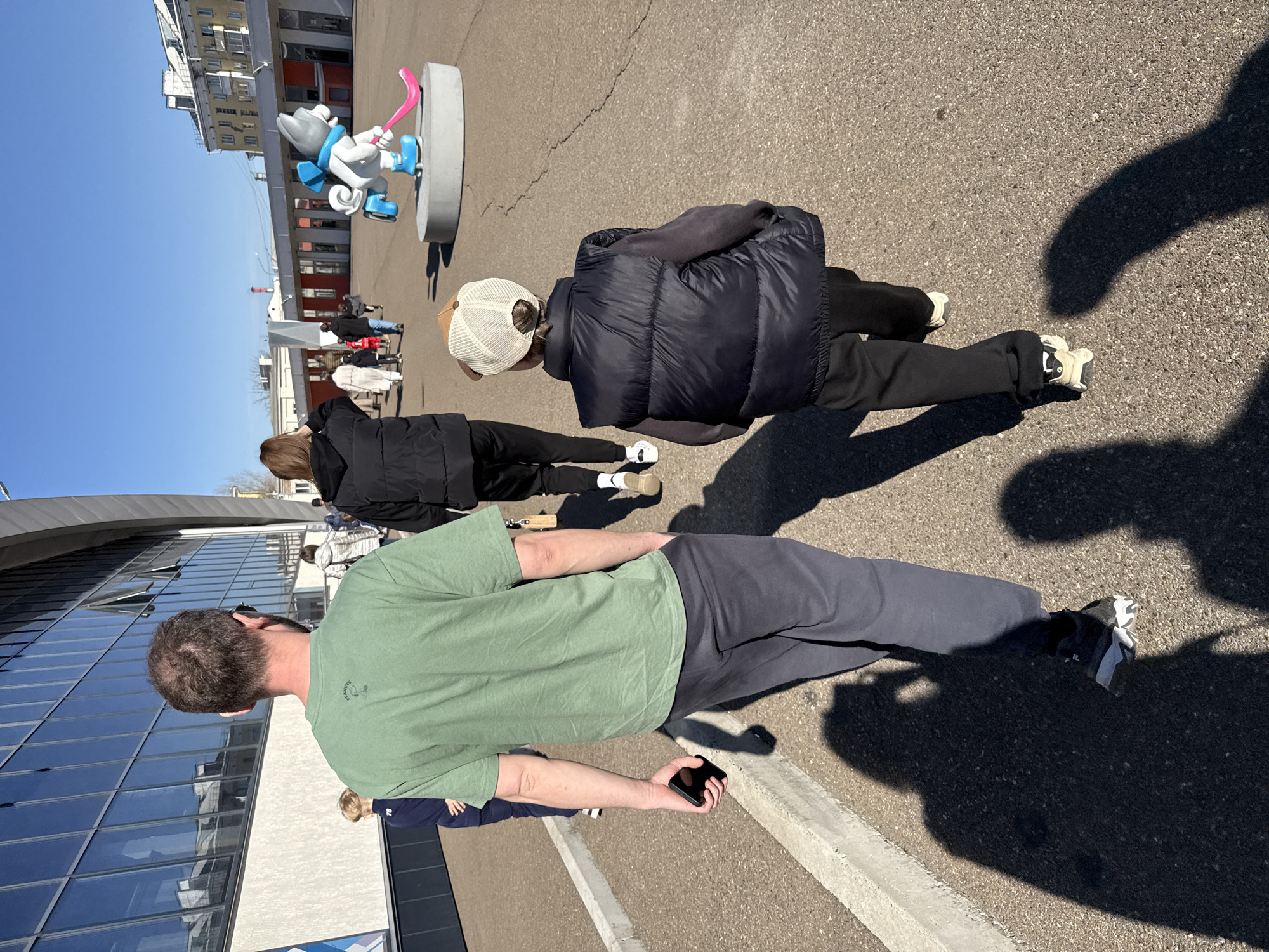 This outdoor image, taken on a sunny day in Krasnoyarsk, Russia, captures several people walking near a sports facility. The sky is clear blue, and bright sunlight casts long shadows, suggesting midday or early afternoon.
In the foreground, a man with short brown hair is seen from behind, wearing a green t-shirt with a small "PEANUTS" logo on the back and dark grey pants. He holds a black smartphone in his right hand. To his right, another person walks ahead, also viewed from behind, dressed in a black puffer vest, a white mesh cap, and dark pants with white sneakers. Further back and to the left, a third individual in a black jacket and dark pants is visible. Other people are indistinctly seen in the background.
A prominent feature in the midground is a large, grey and white cartoonish statue, possibly a mascot, standing on a grey cylindrical base. The figure wears a blue scarf and blue ice skates, holding a pink hockey stick, strongly resembling Uka, the mascot of the 2019 Winter Universiade held in Krasnoyarsk.
On the left side of the image, a modern building with a curved, reflective glass facade suggests a sports complex or arena. In the background, older, multi-story urban buildings with some orange-red elements line the street. The ground is paved asphalt. The scene depicts a casual public setting, likely near a sports venue, with people going about their day.