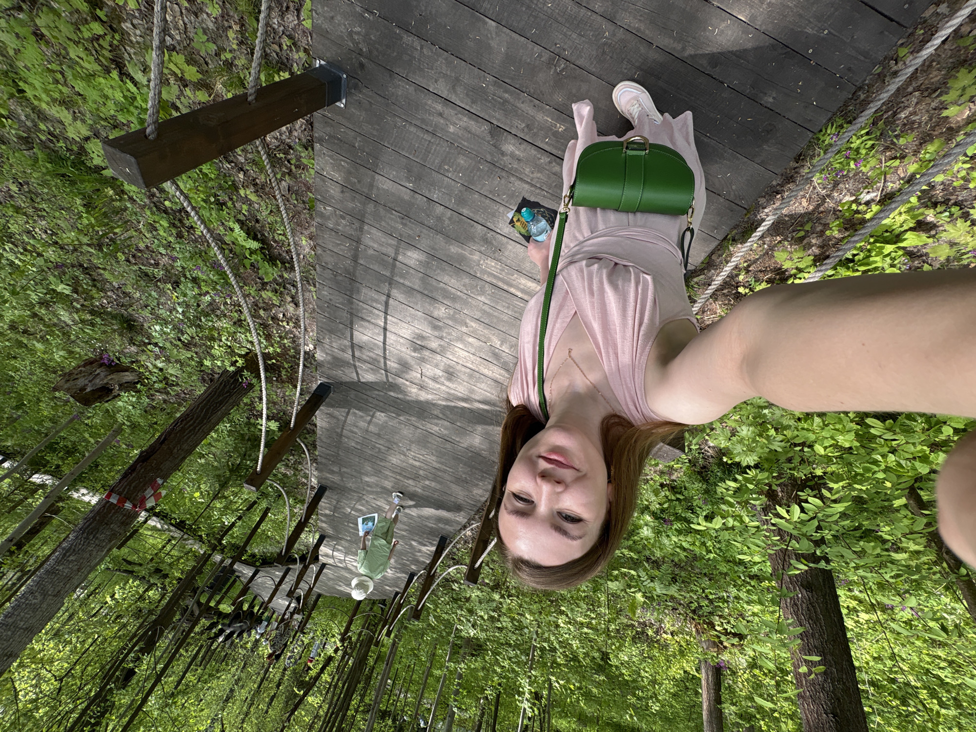 A young woman is taking a selfie on a wooden walkway in the city of Izhevsk, Russia. She is wearing a light pink dress and sneakers, with a green purse over her shoulder. She is holding a bottle of water and posing for the photo. The walkway is elevated and surrounded by trees and foliage. Another person can be seen further down the walkway.