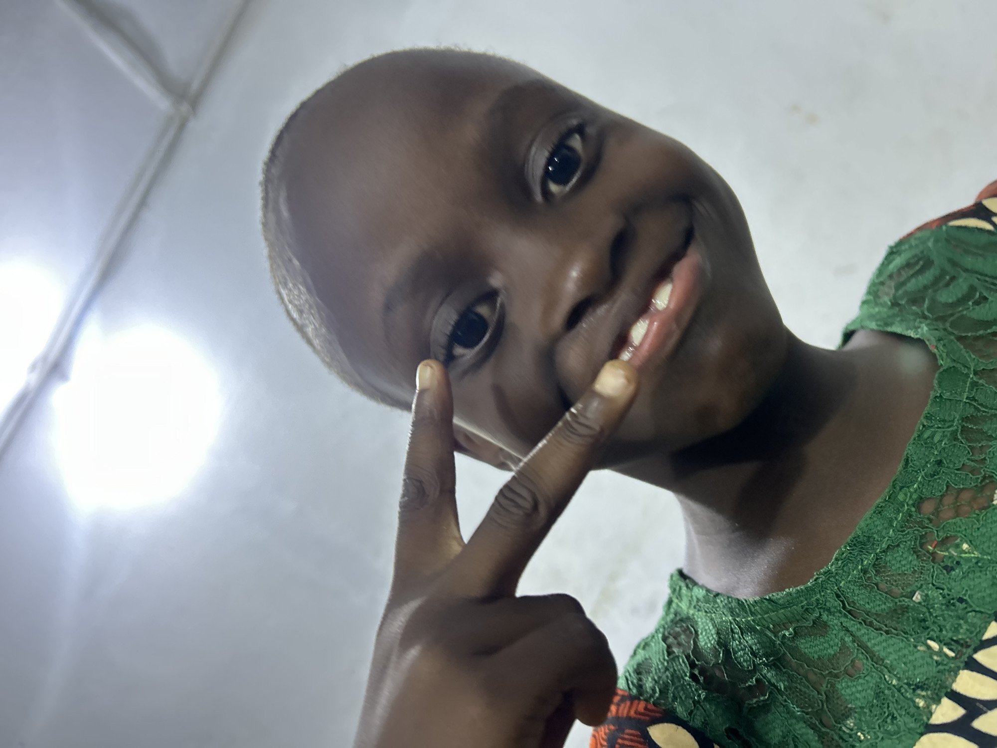 A young individual with dark skin, possibly a child, is smiling and holding up a hand in a "peace" or "victory" sign. They appear to be indoors. The individual is wearing a garment with a green lace collar. The background is a plain white wall, which is brightly lit with an undefined light source, possibly a flash. The location is Unknown, Unknown.