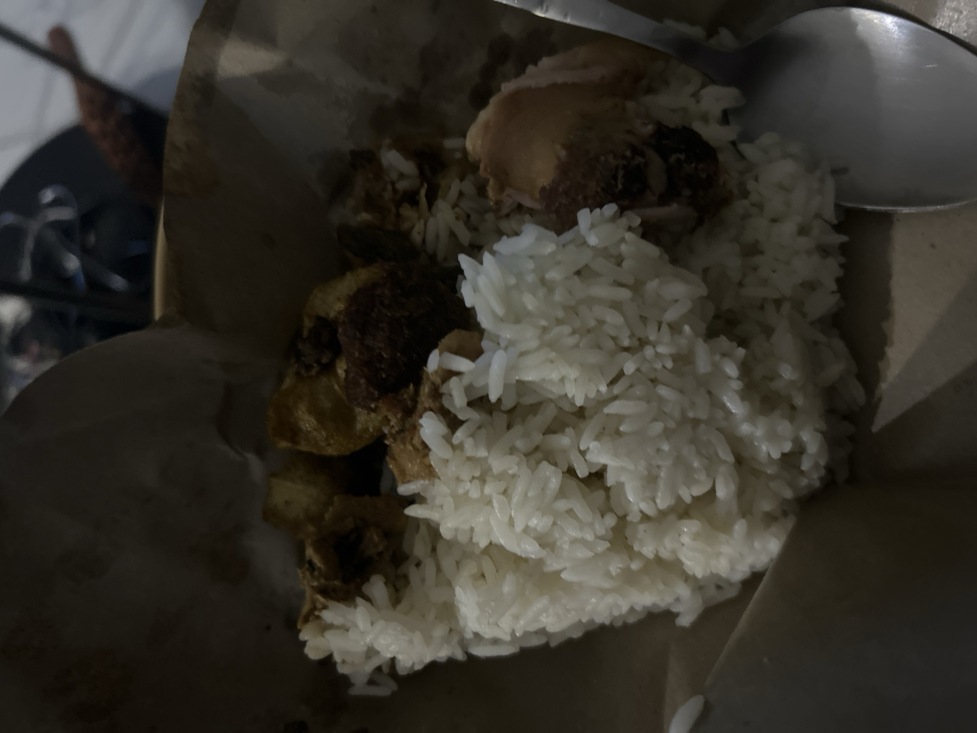 A close-up, slightly blurry, overhead shot shows a portion of white rice served in a brown paper wrapper, with a spoon to the right. Several pieces of cooked chicken and some other food items, possibly vegetables or potatoes, are visible mixed with the rice. The lighting is dim, suggesting an indoor or evening setting. The image's contents are suggestive of a takeaway meal. The context of Sukawati, Indonesia, implies this meal might be traditional Indonesian cuisine.