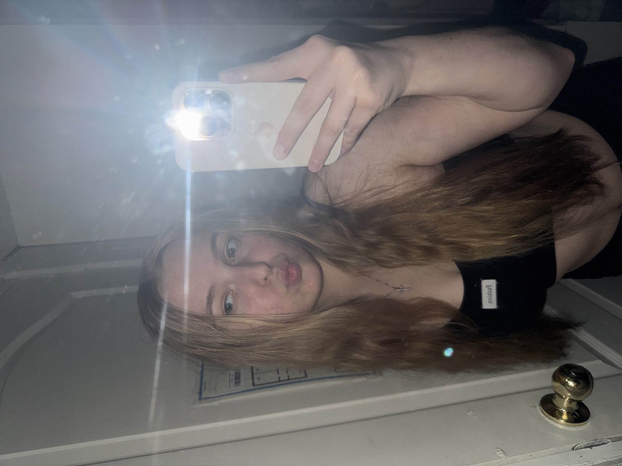 A young woman is depicted in a mirror selfie, posing with pursed lips and looking to her right. She has long, light brown hair and fair skin. She is wearing a black crop top with a small, inverted white label that reads "noideЯ" and a silver-colored necklace featuring a small cross pendant. Her left hand holds a light-colored smartphone, likely an iPhone, whose active flash creates a strong light source and prominent lens flare, partially obscuring the upper right portion of the image. The background, reflected in the mirror, shows an indoor environment featuring a white door with a brass-colored doorknob on the left. Behind the woman's head, a white rectangular object with dark lines, possibly a diagram or plan, is visible on the wall. The scene is dimly lit, with the phone's flash being the primary light source.