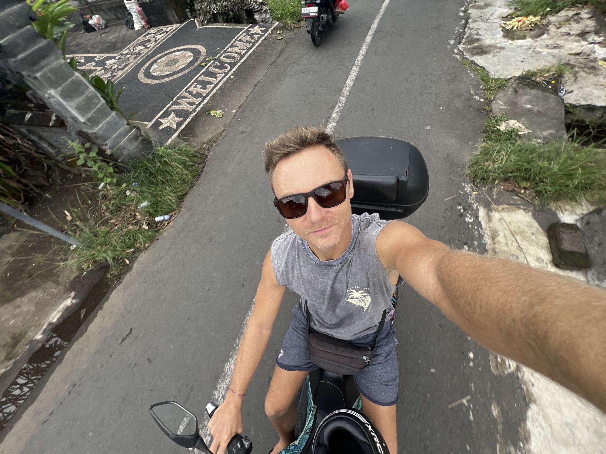 A man is riding a scooter on a road in Sukawati, Indonesia. The image is a selfie taken by the man, who is wearing sunglasses and a gray tank top and shorts. He has a black bag slung across his chest and a helmet near the scooter's seat. Behind him is a black storage container mounted on the scooter. The road has a white dividing line. On the left side of the road, there's a decorative stone mat with the word "WELCOME" inscribed on it. Another scooter is visible further up the road. Vegetation and concrete structures border the road on both sides. The overall lighting suggests it is daytime.