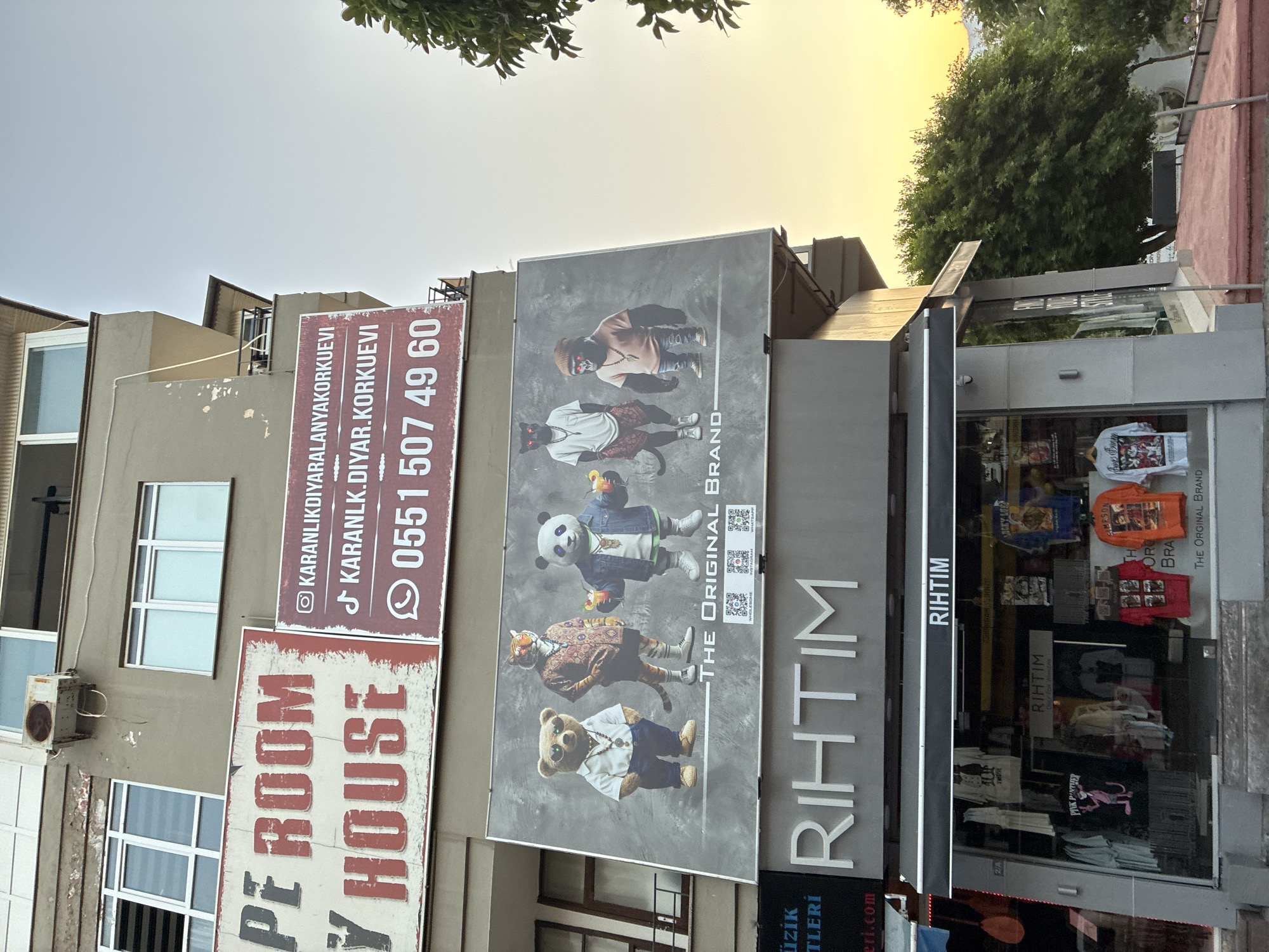 The image captures a street-level view of a building in Alanya, Turkey, featuring a large billboard advertising a business. The billboard displays a whimsical design with anthropomorphic animals dressed in trendy clothing, including a bear, a tiger, a panda, a black panther, and a gorilla. The text "THE ORIGINAL BRAND" is prominently featured below the animal figures. Above this billboard, another sign reads "PE ROOM HOUSE" in large, distressed red lettering. To the right of "PE ROOM HOUSE," a smaller sign displays contact information, including social media handles and a phone number.
Below the billboard, the storefront of a shop named "RIHTIM" is visible. The shop's interior, visible through large glass windows, displays various clothing items, primarily t-shirts, with some featuring graphics like "Pink Panther" and other designs. A black awning extends over the shop entrance. The overall scene is illuminated by the warm glow of what appears to be a sunset, casting a soft light on the building and the surrounding greenery. The architectural style suggests an urban environment, likely a commercial street.