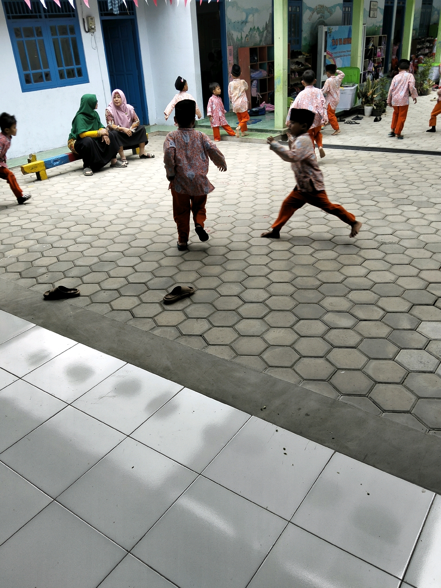 In Krajan Lor, Indonesia, a group of children in traditional clothing are playing on a paved courtyard. The courtyard is covered with hexagonal gray tiles. In the foreground, two pairs of dark-colored sandals are placed on the pavement. Behind them, a group of children dressed in colorful shirts and pants are running and playing. Some of the children are wearing small caps. Two women, one in a green hijab and the other in a pink hijab, are sitting on a yellow and blue seesaw in the background. The building in the background has white walls and blue windows. The overall scene depicts a lively outdoor play area for children.
