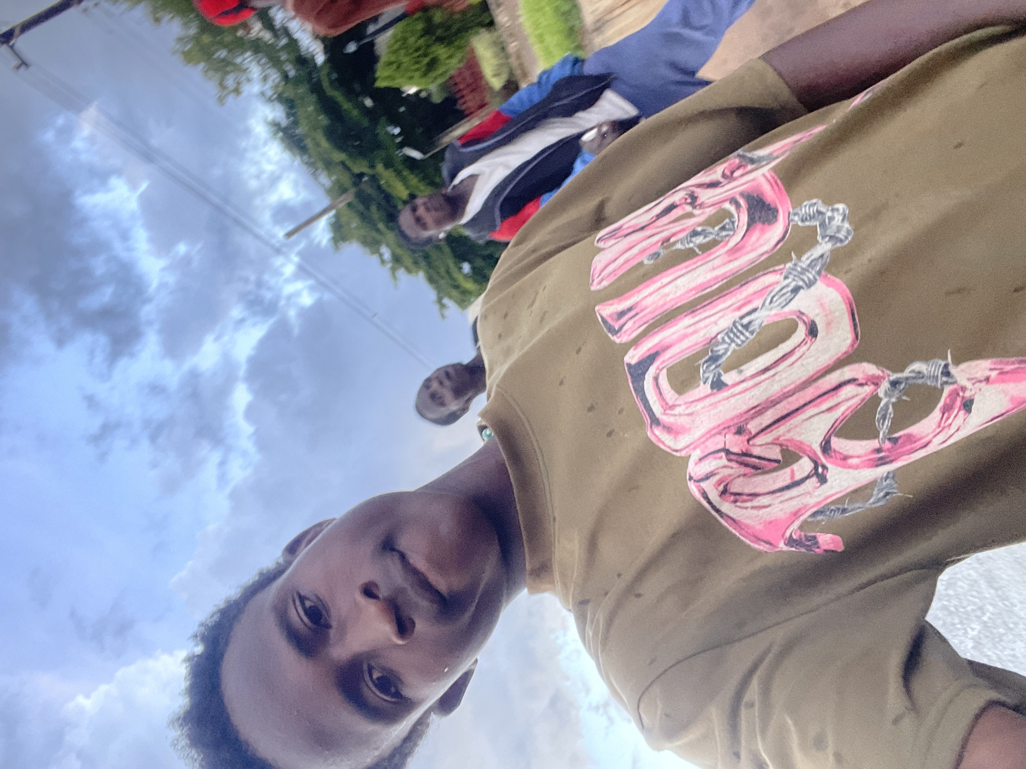In a low-angle, close-up selfie, a young Black male faces the camera, his head tilted slightly. He is wearing a khaki-colored t-shirt with a large, stylized pink graphic featuring the word "BOU" entwined with barbed wire. In the background, three other individuals are visible. To the left of the main subject, a Black male is seen from the chest up, looking away from the camera. Behind him, another Black male, also seen from the chest up, is walking away, wearing a dark jacket with red and blue accents over a white shirt. Further to the right and partially obscured, a person wearing a red hat and a brown shirt is visible. The setting appears to be outdoors, with a cloudy sky overhead and trees and vegetation lining a paved area in the background. The overall scene suggests a casual outdoor gathering or stroll. The image was taken in Jos, Nigeria.