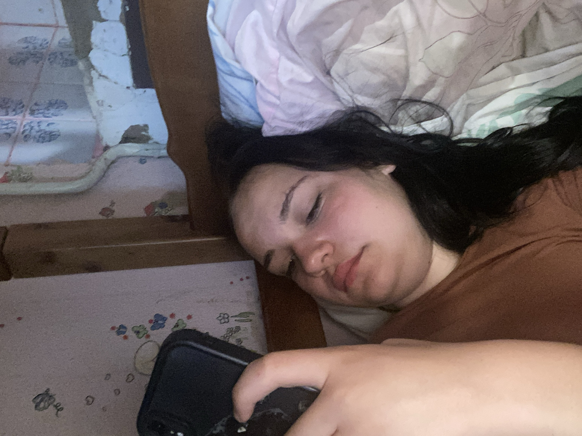 A young person with dark hair lies in bed, holding a black smartphone. The person is wearing a brown shirt. The background consists of a wall with pink wallpaper featuring butterflies and hearts. A wooden bed frame is visible on the right side of the image, and a portion of a blue and pink comforter is present. The lighting is dim.
