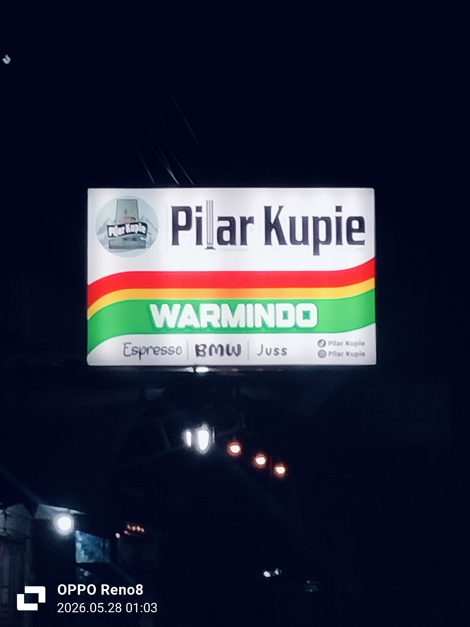 At night, a sign illuminated above a shop or building reads "Pilar Kupie" with a logo of a building. Below the name is "WARMINDO" and "Espresso BMW Juss". There is a green, yellow and red stripe between the store name and warmindo. The image was taken with an OPPO Reno8, indicated by the watermark in the lower left corner, time-stamped 2026.05.28 01:03. The location is likely Cempeudak, Indonesia, based on the implied context of the brand name. The background is dark and out of focus, with indistinct street lights.