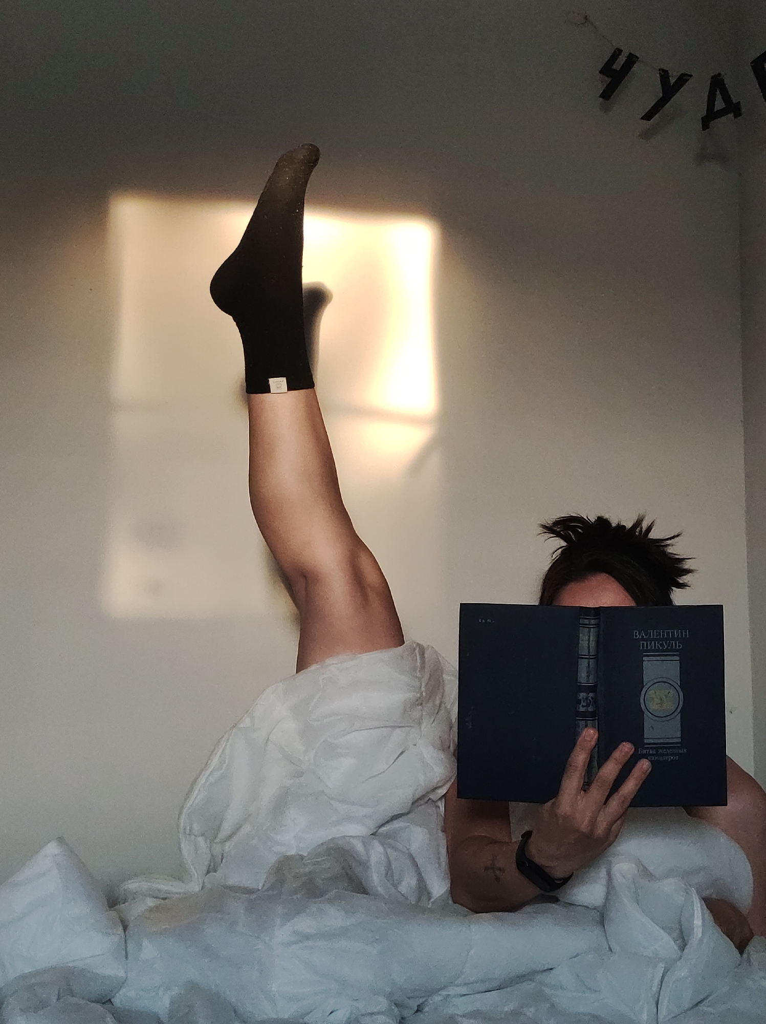 A person is lying in a bed with white sheets and blankets, reading a book titled "Валентин Пикуль" (Valentin Pikul). The person's left leg is raised vertically, with a black sock on the foot. A section of a wall with a window is visible behind the person, and the word "ЧУД" (CHUD) is hanging on the wall. The location is an indoor setting in the city of Unknown, Unknown.
