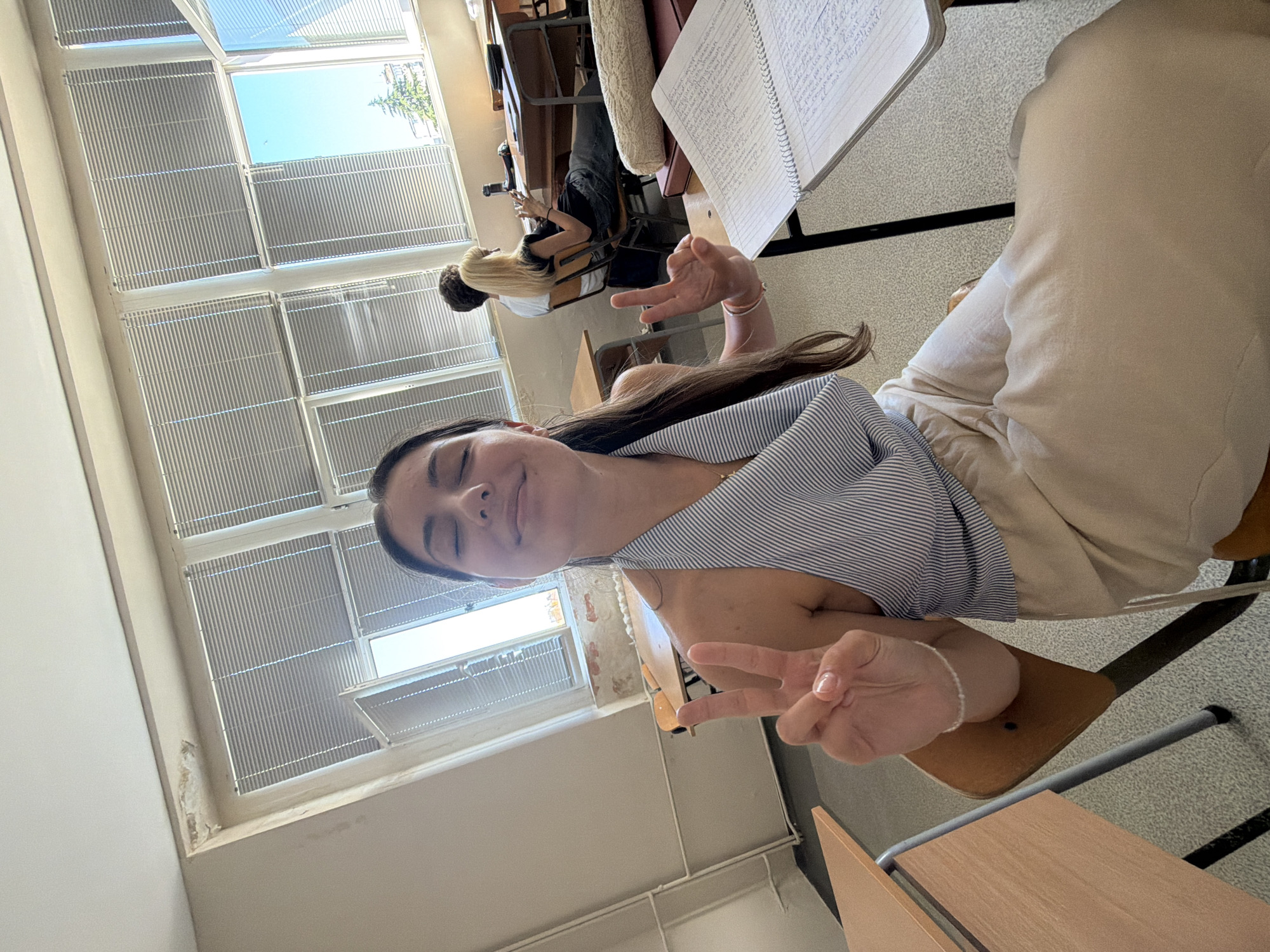 A young woman with her eyes closed and a smile on her face, sitting in a classroom in Burgas, Bulgaria, poses for a selfie, holding up two fingers in a peace sign with her left hand and the index finger and thumb in an 'okay' gesture with her right hand. She is wearing a striped halter top and light-colored trousers. The classroom environment includes desks, chairs, and large windows with blinds. Two other individuals, a man and a woman, are visible in the background, seated at desks. The windows offer a view of trees and a building, indicating an outdoor setting beyond the classroom. The room appears somewhat dated, with visible wear and tear on the walls near the ceiling.