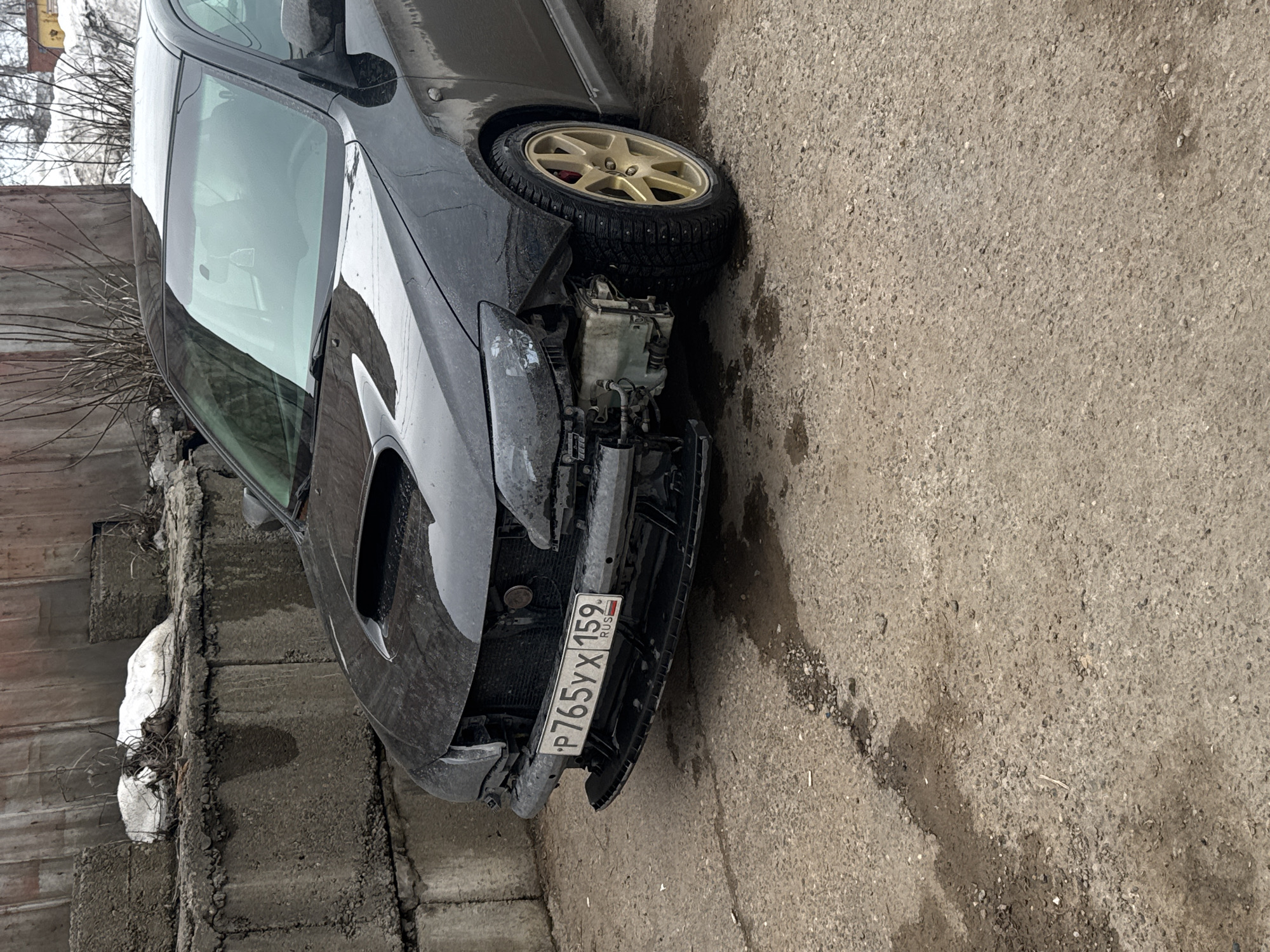 A dark-colored Subaru Impreza, bearing Russian license plate P765YX 159, registered in Perm Krai (which includes the city of Chaikovsky), is parked on a rough concrete surface. The vehicle has sustained significant frontal damage, with the front bumper partially detached and hanging. The front grille is missing, and the driver's side headlight assembly is damaged. Exposed mechanical components, possibly part of the cooling or air conditioning system, are visible on the passenger side of the front bay. The front passenger-side wheel features a gold-colored multi-spoke rim fitted with a studded winter tire, and a red brake caliper is visible behind it. In the background, a retaining wall constructed from large concrete blocks is present, topped by a weathered reddish-brown corrugated metal structure. Sparse, bare tree branches and patches of white material, likely snow or debris, are visible behind the wall. The ground around the vehicle shows scattered small stones and damp patches. No individuals are present in the scene.