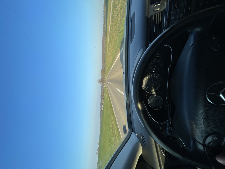 The image presents a first-person perspective from within a moving Mercedes-Benz vehicle, traveling on a rural asphalt road under a clear, bright blue sky in the vicinity of Weißenhorn, Germany. The time appears to be late afternoon or early evening, indicated by the low sun angle and soft lighting.
In the immediate foreground, the black leather steering wheel, emblazoned with the Mercedes-Benz logo, occupies the lower central portion of the frame. A driver's left hand is partially visible gripping the lower left section of the steering wheel. Behind the wheel, the vehicle's instrument cluster is visible, showing a speedometer needle resting near 90 km/h, a digital readout confirming 91.0 km/h, an external temperature of 23.0°C, and a remaining fuel range of 260 km. The dashboard is dark-colored, and air vents are visible on the passenger side. The windshield displays some minor dust or debris.
The two-lane road stretches straight ahead, marked by solid white lines on the shoulders and a dashed white center line, eventually curving slightly to the right in the distance. Lush green fields flank both sides of the roadway. On the left side, a wooden hunting blind or deer stand is visible in the field, with a small cluster of houses, indicating a hamlet or village, further on the horizon. The right side features additional green fields bordered by a line of leafless deciduous trees, suggesting a late autumn or winter season. A solitary, bare tree stands prominently near the road's vanishing point in the background. No other vehicles or individuals are discernibly present on the road.